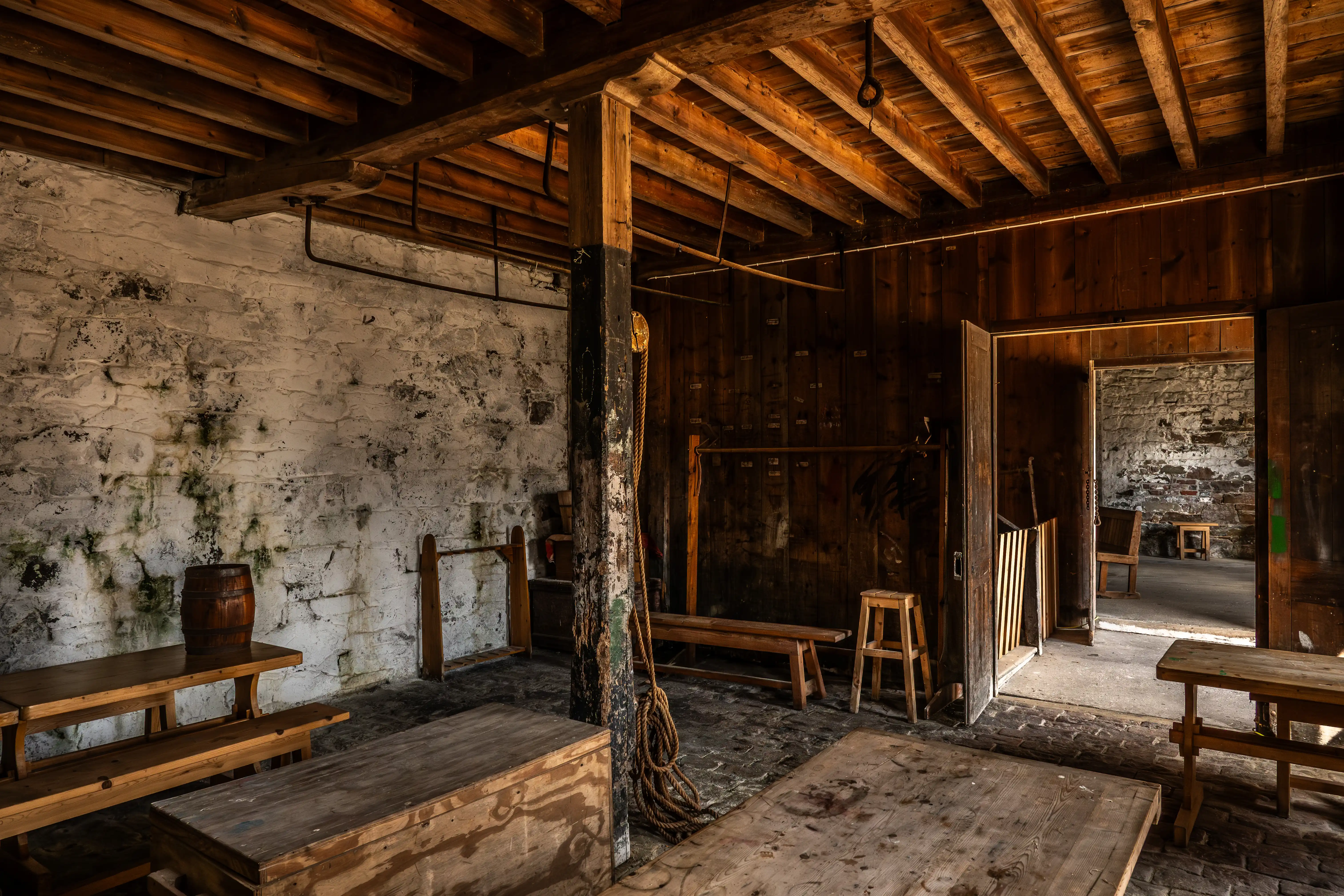 Interior of a dimly lit, rustic building with wooden ceiling beams, rough white walls, wooden tables and benches, a wooden post, and an open doorway revealing another stone-walled room.
