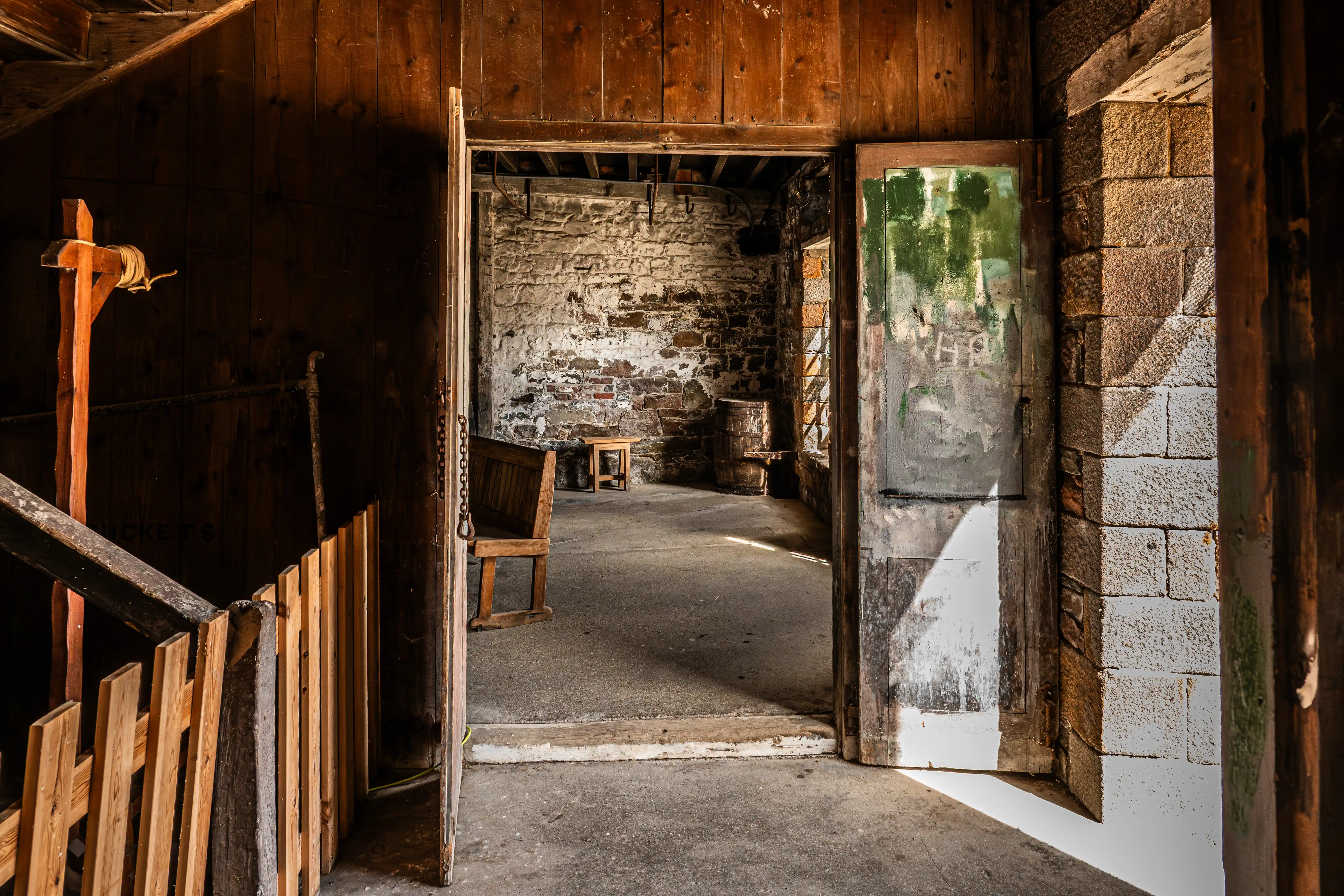 Interior of an old rustic building with a wooden staircase and fence on the left, opening to a stone-walled room containing a wooden bench, a small stool, and a barrel. Sunlight illuminates the floor from the right.