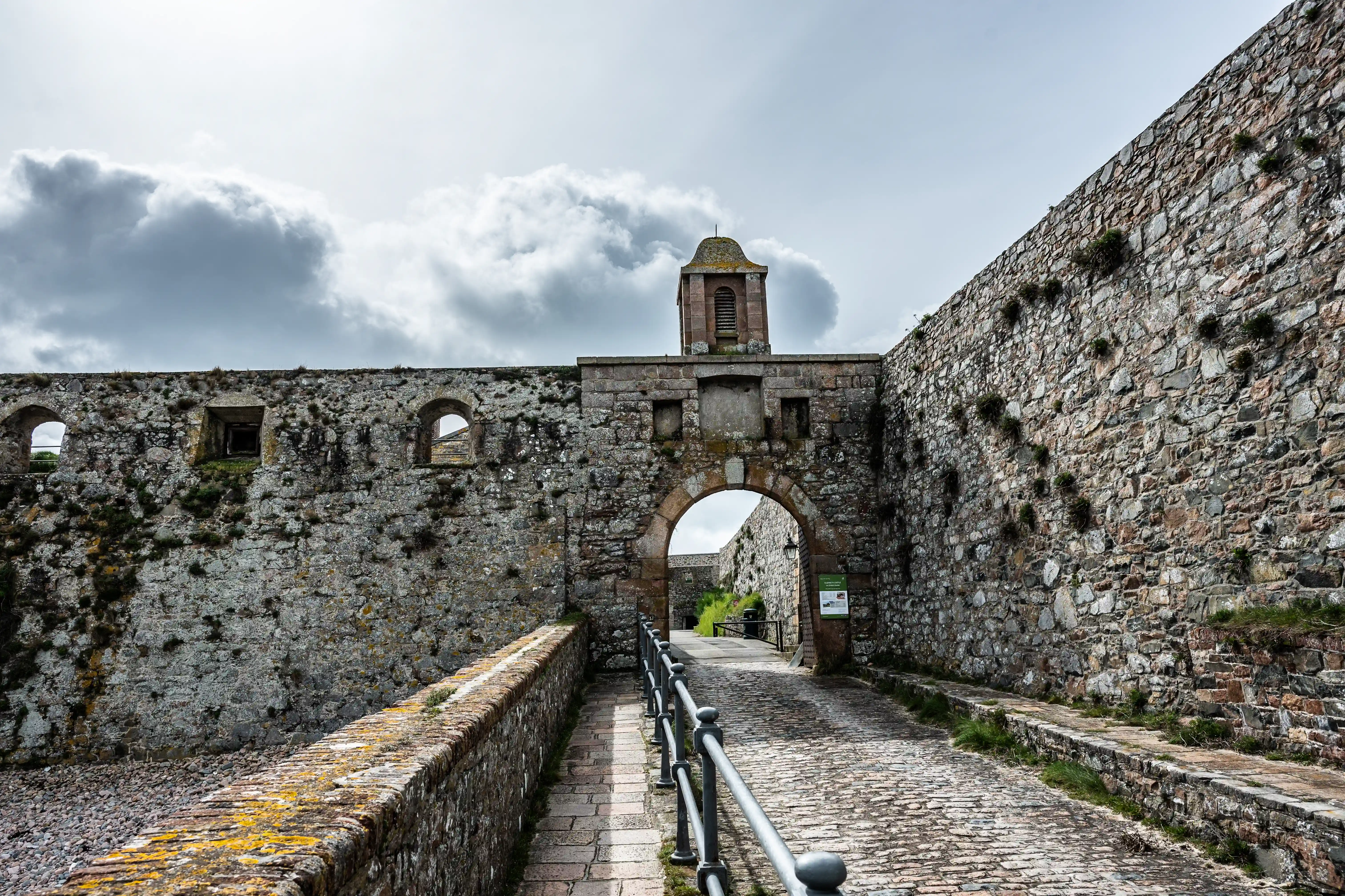 Stone archway entrance to a historic fortress, with a cobbled pathway and flanking stone walls under a dramatic cloudy sky.