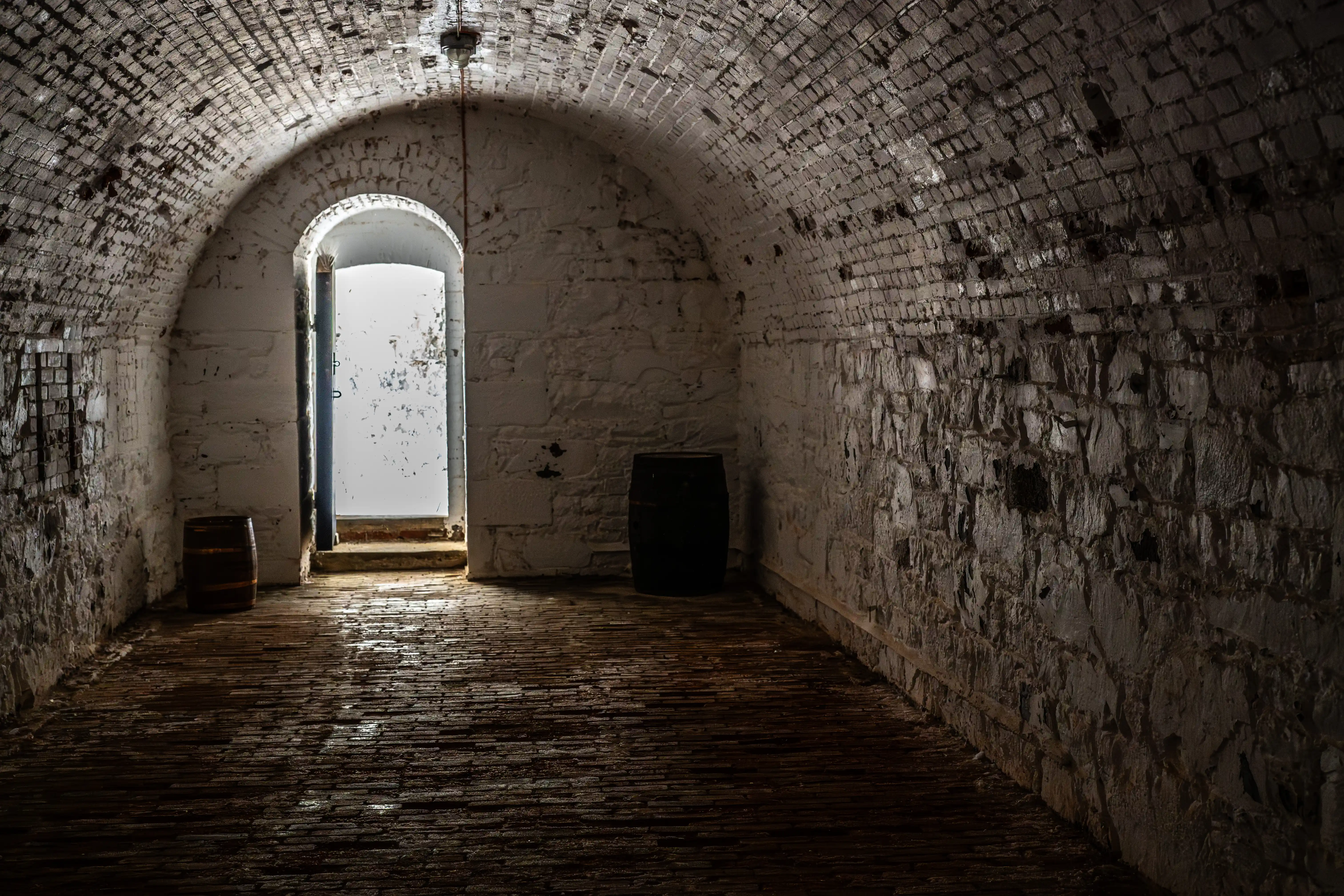 Interior view of a dark, arched brick corridor with a wet brick floor. Two barrels are visible, and a bright, open doorway is at the end of the passage.
