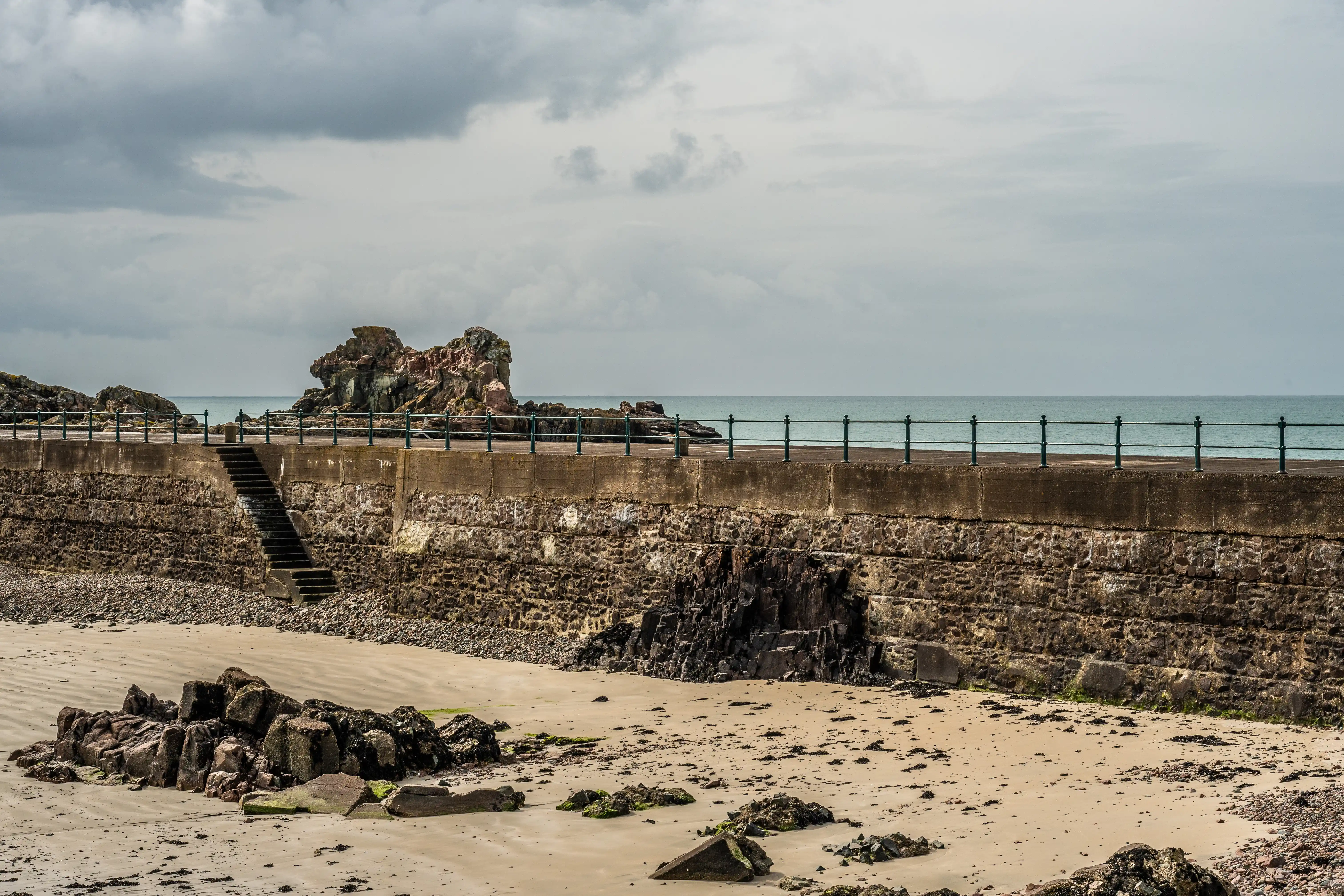 A wide shot of a stone harbour wall on a beach under a cloudy sky, with the sea in the background.