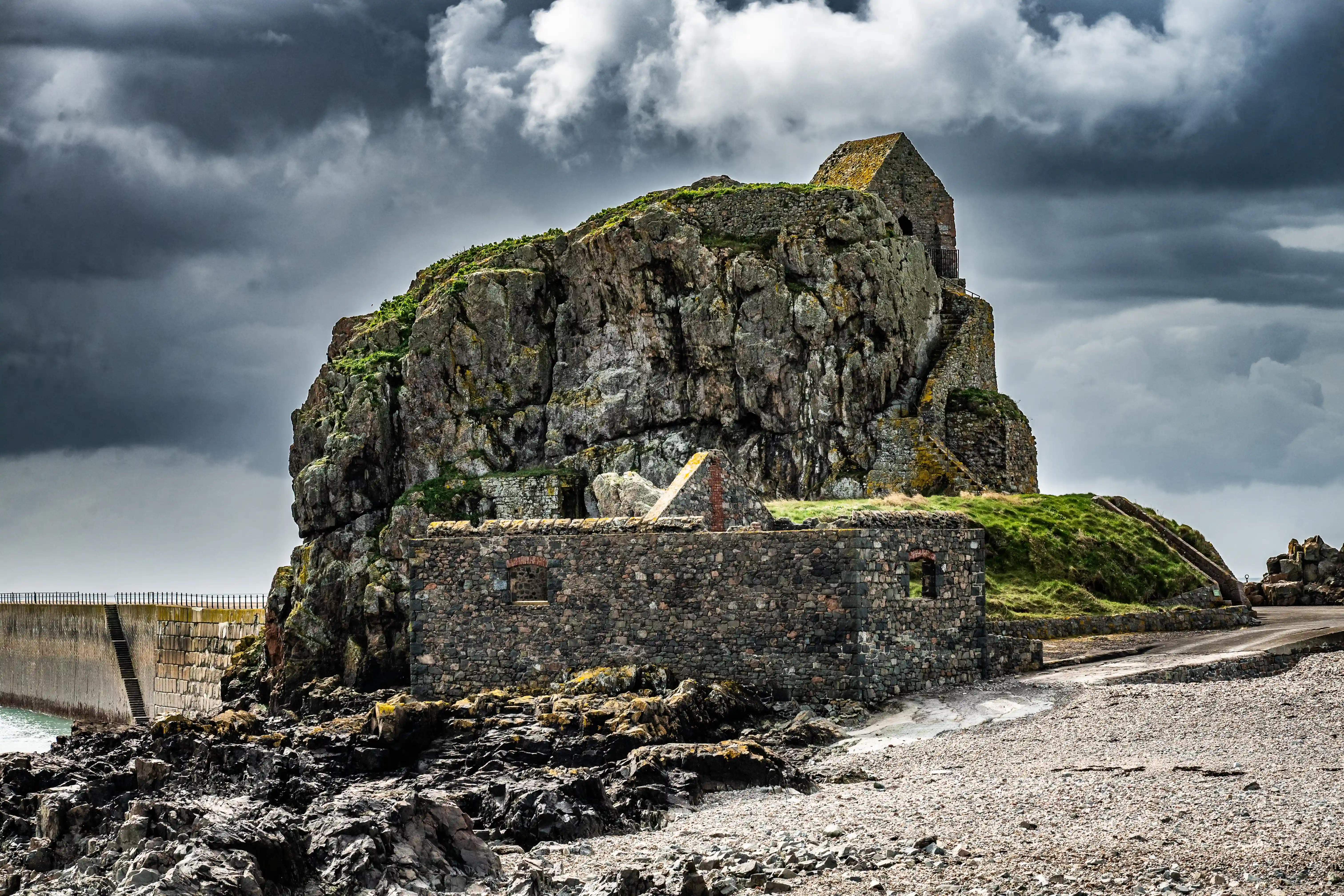 A wide shot of a rocky island with derelict stone buildings, including a prominent structure with a pointed roof at the summit. A substantial sea wall is visible on the left, and a rough path leads towards the structures from a pebbled shore. The sky is heavily clouded with dark, moody tones.