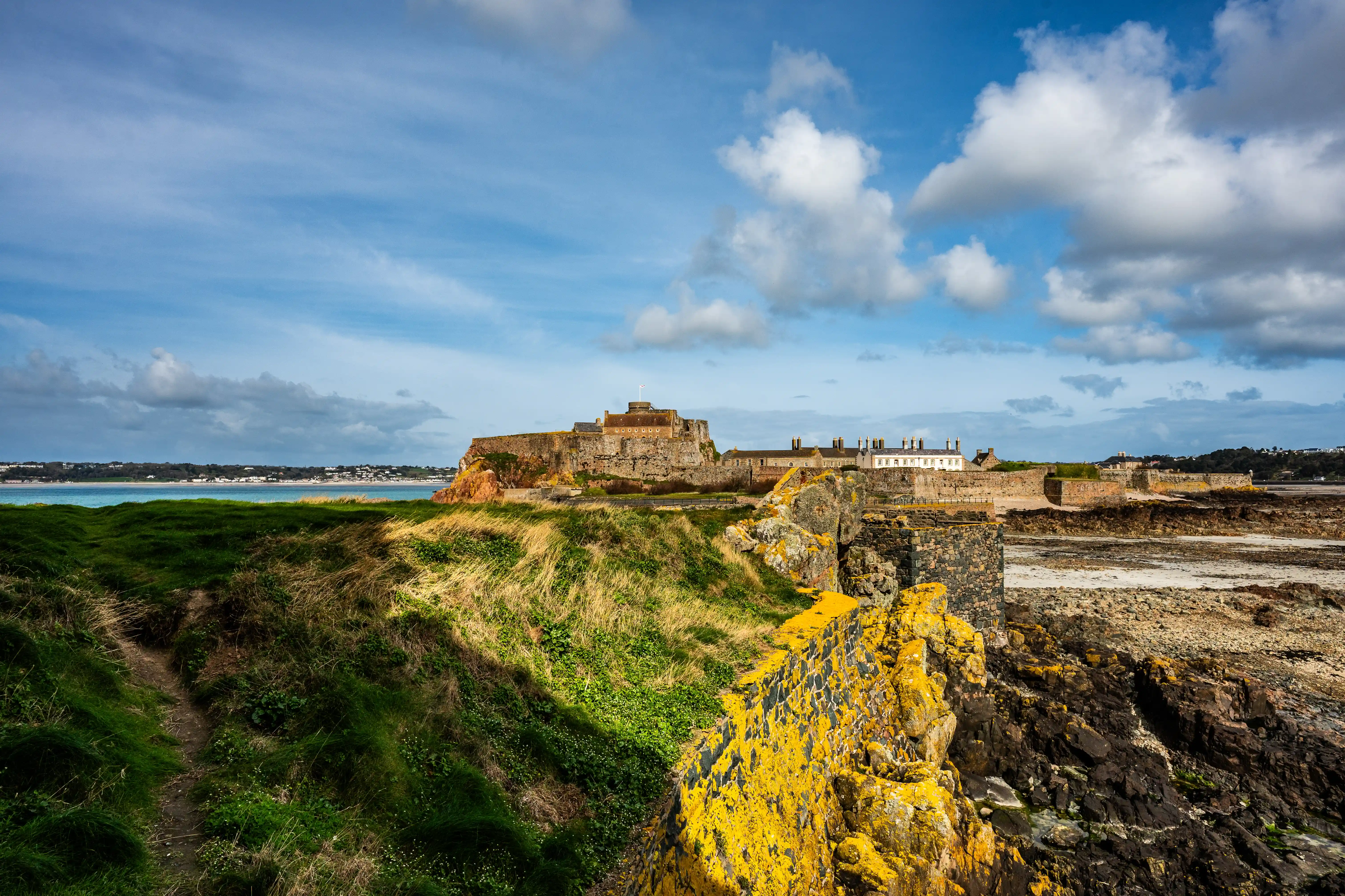 A panoramic view of a historic stone castle on a cliff, with green grassy slopes in the foreground and a blue sky with white clouds above.