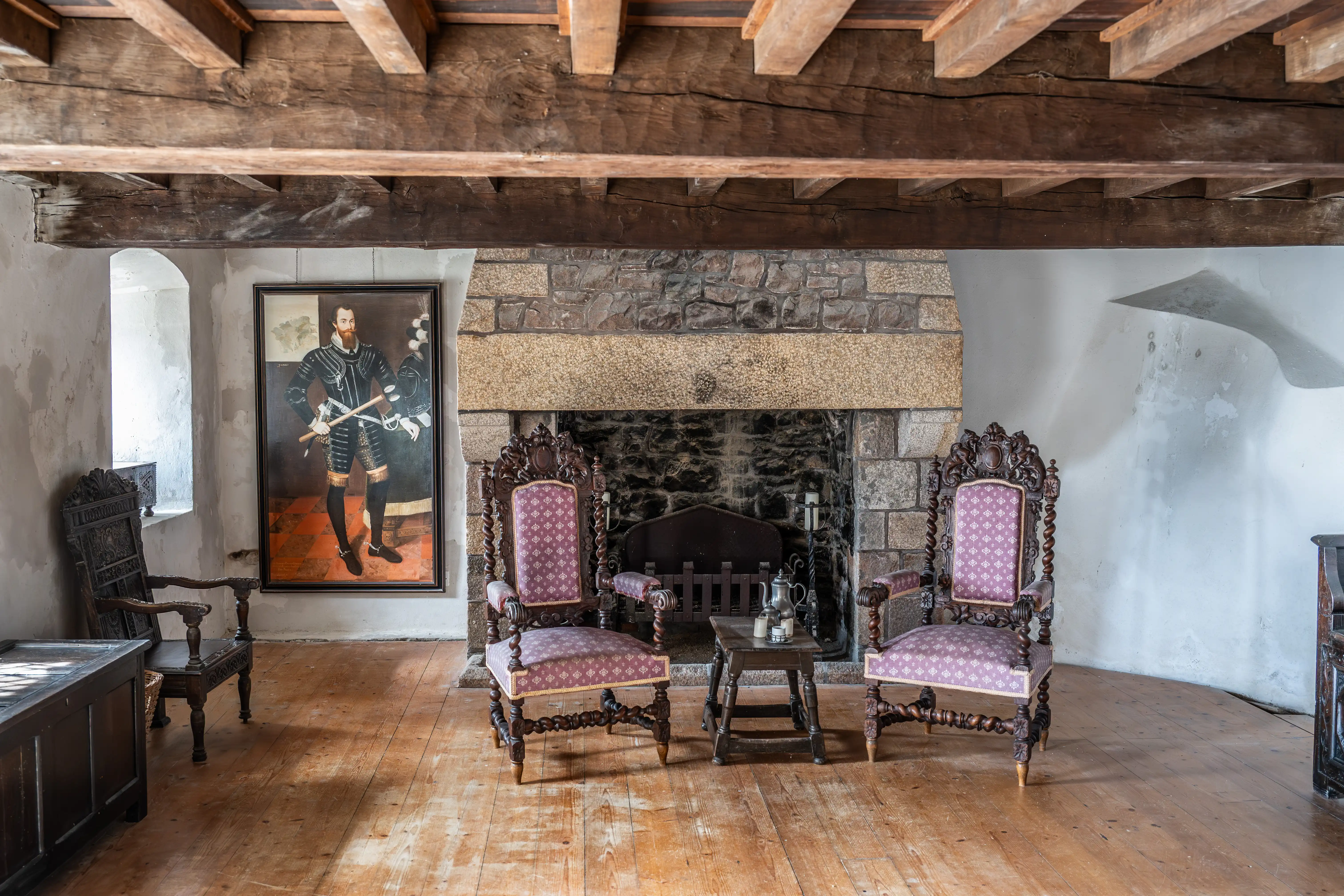 Interior view of a historic room with a stone fireplace, two purple upholstered armchairs, a portrait of a man, and wooden ceiling beams.