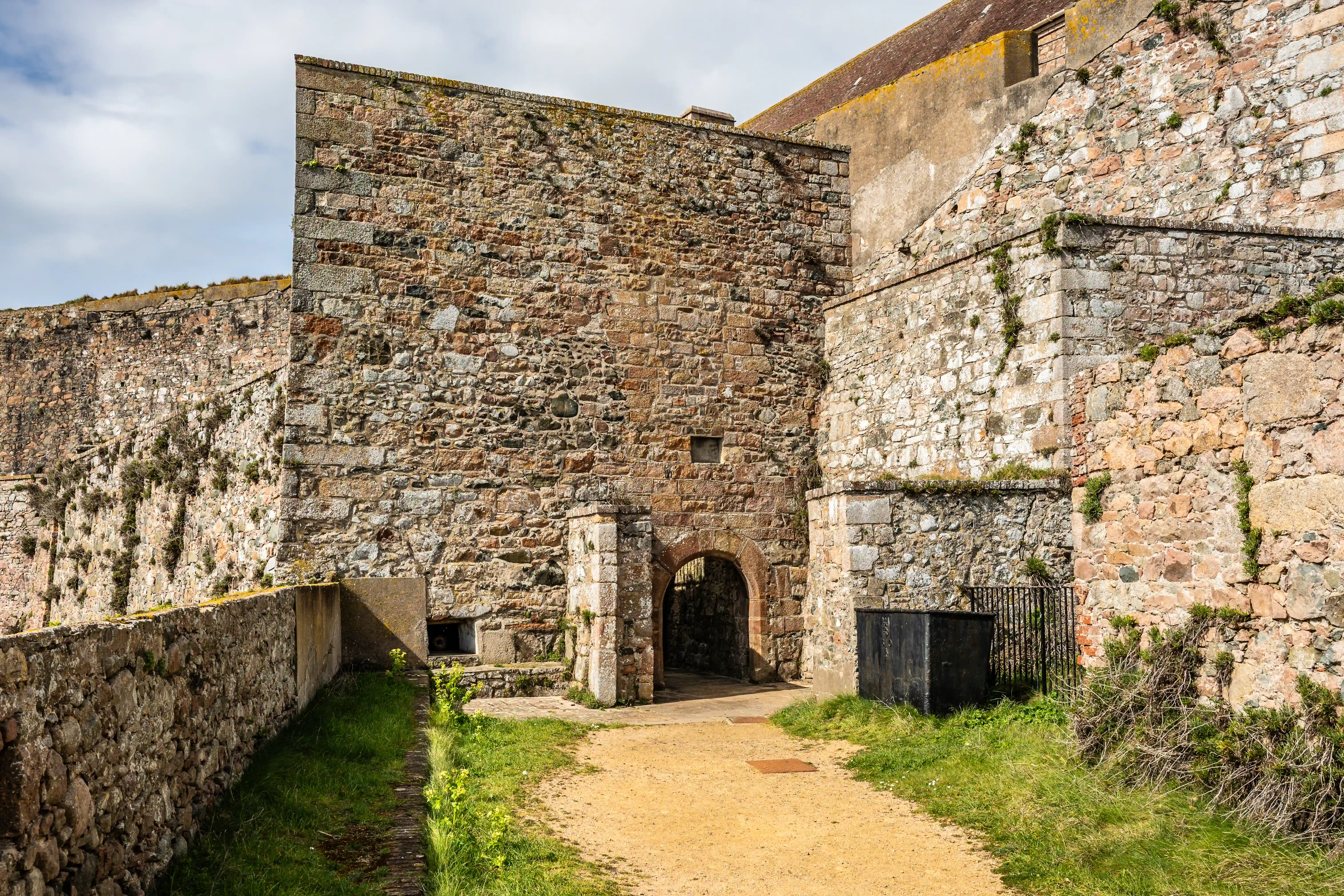 A sandy path approaches an arched stone gateway within a weathered stone fortress. The walls are constructed of rough-hewn stones, with moss and small plants growing in crevices. A low stone wall borders the left side of the path, and a dark metal bin stands to the right of the archway. The sky above is partly cloudy.