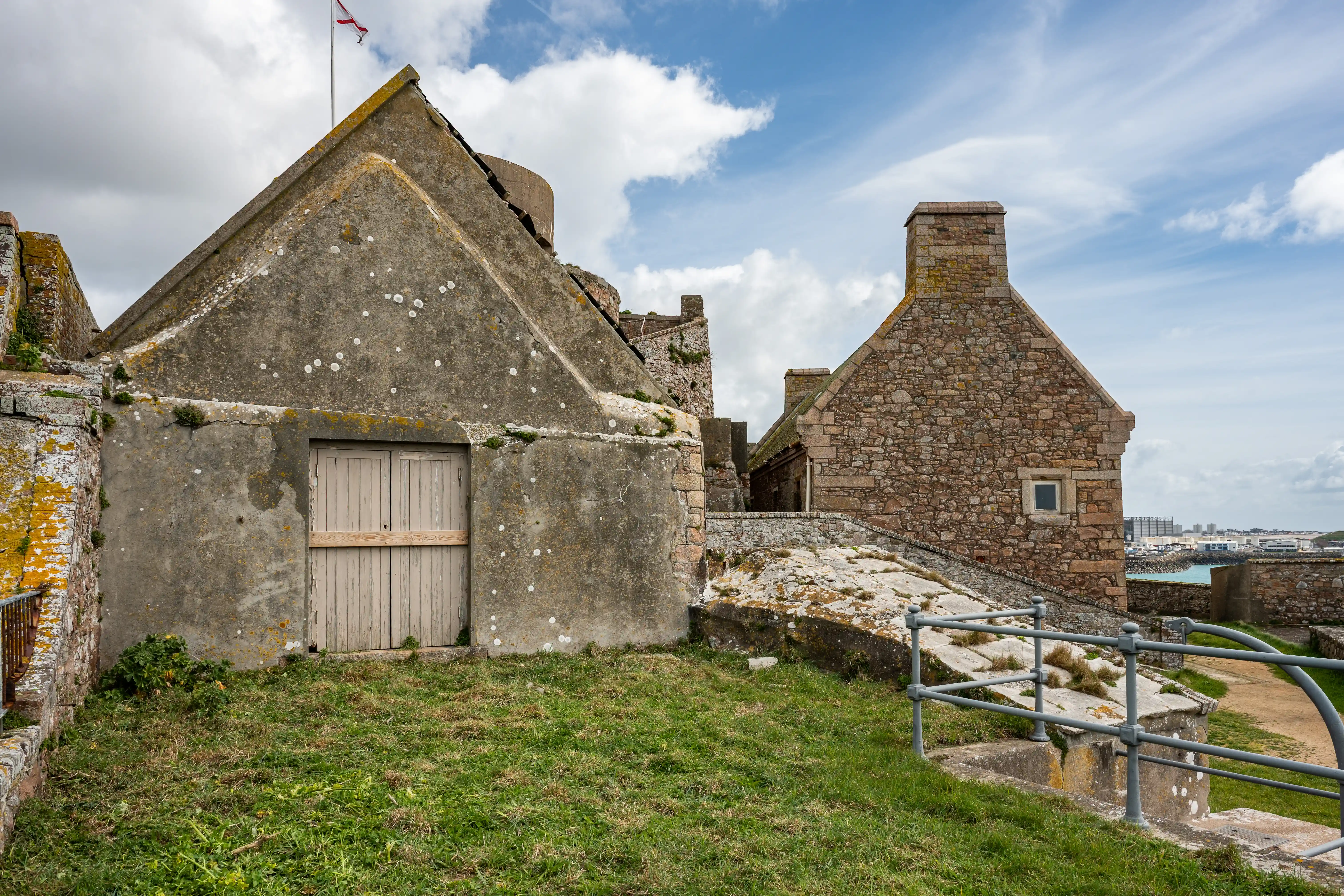  A weathered concrete building with wooden doors and a pitched roof stands in the foreground, to the left of a more traditional stone building with a chimney, set against a backdrop of sky and distant buildings.