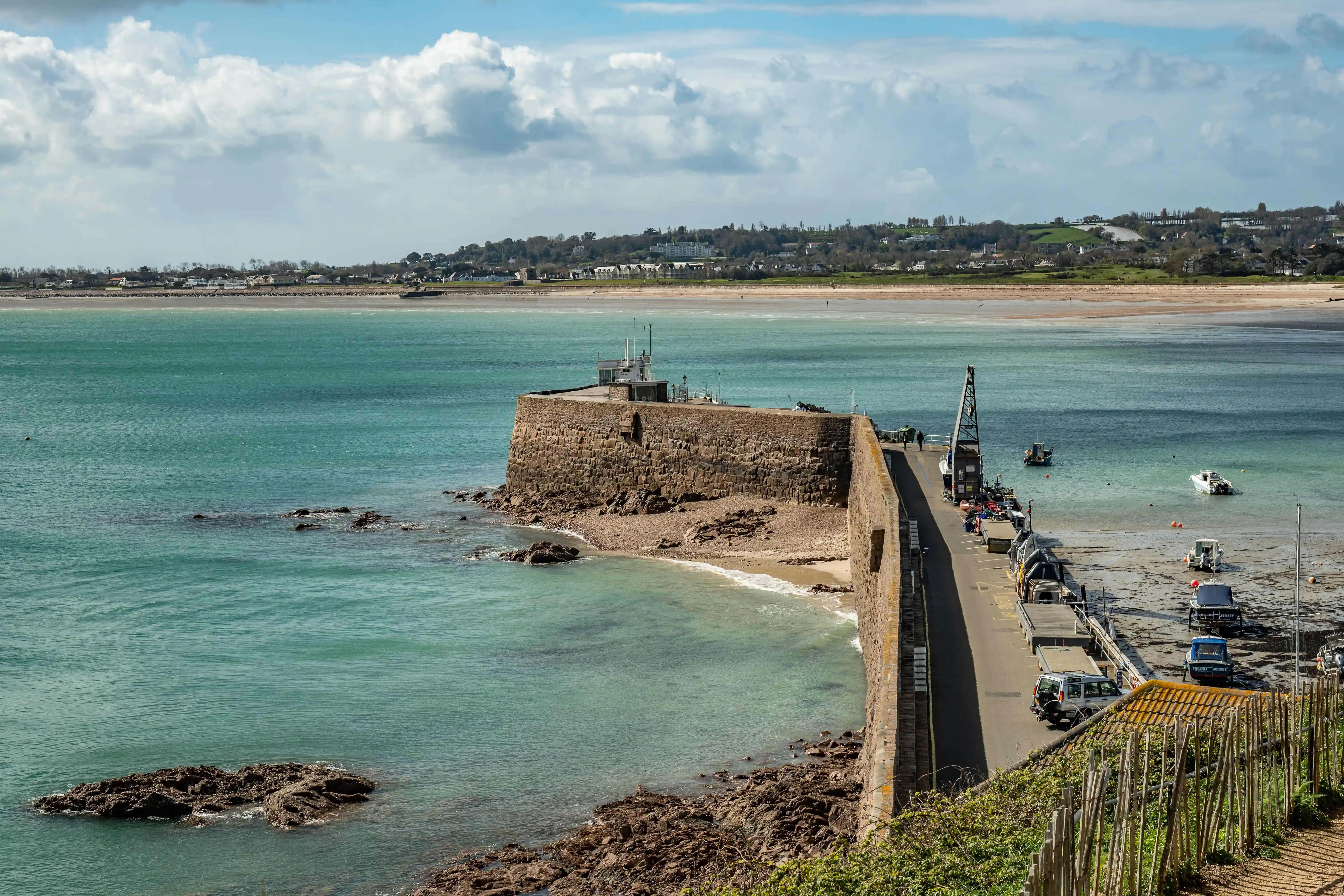 A wide shot of a harbour with a large, weathered stone breakwater curving into clear turquoise water. Several small boats are visible in the bay. A beach with gentle waves can be seen inside the breakwater, and a coastal town is in the distance under a blue sky with scattered clouds.