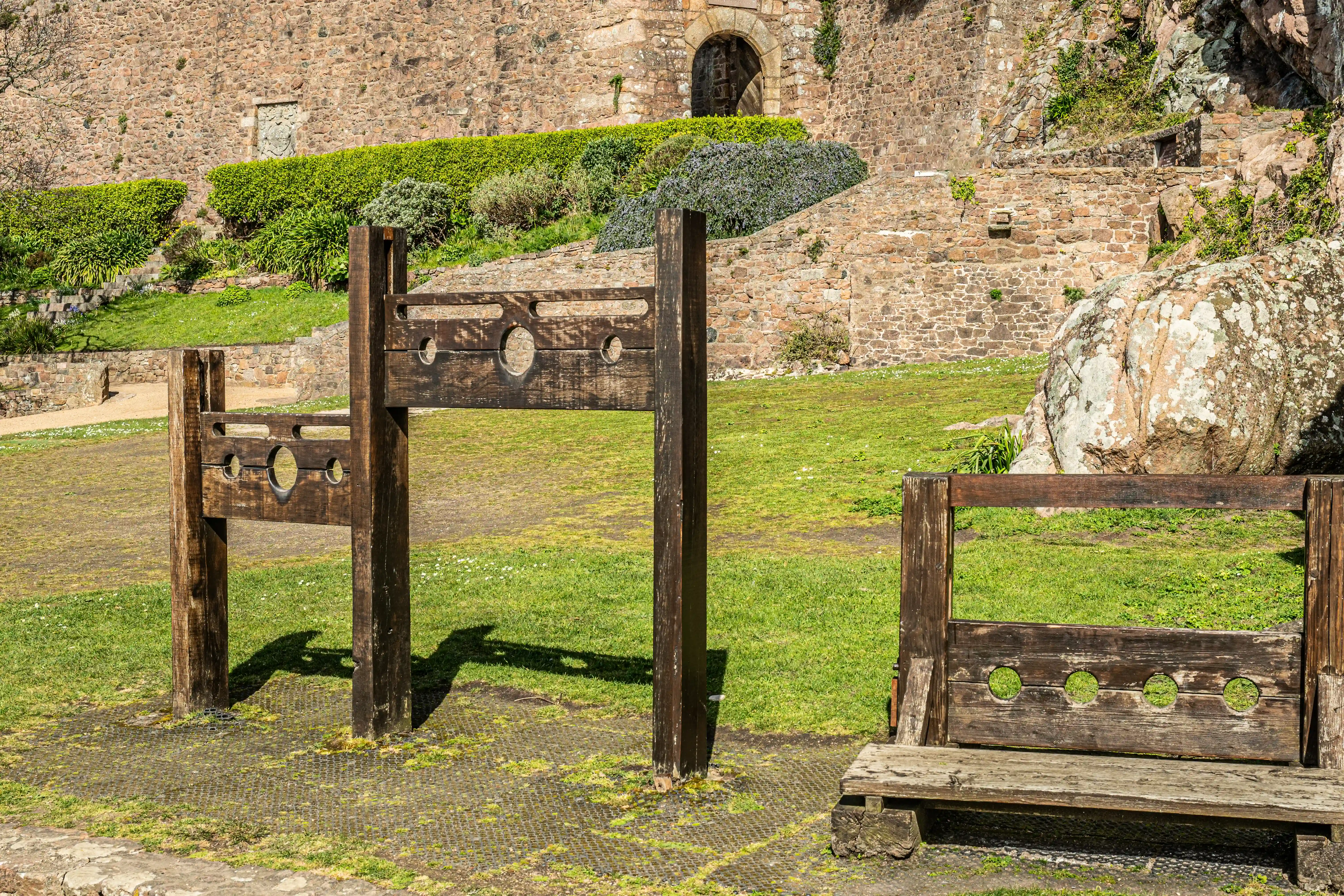 Wooden stocks with openings for hands and feet, situated on a grassy area in front of a medieval stone castle wall with an arched entrance.
