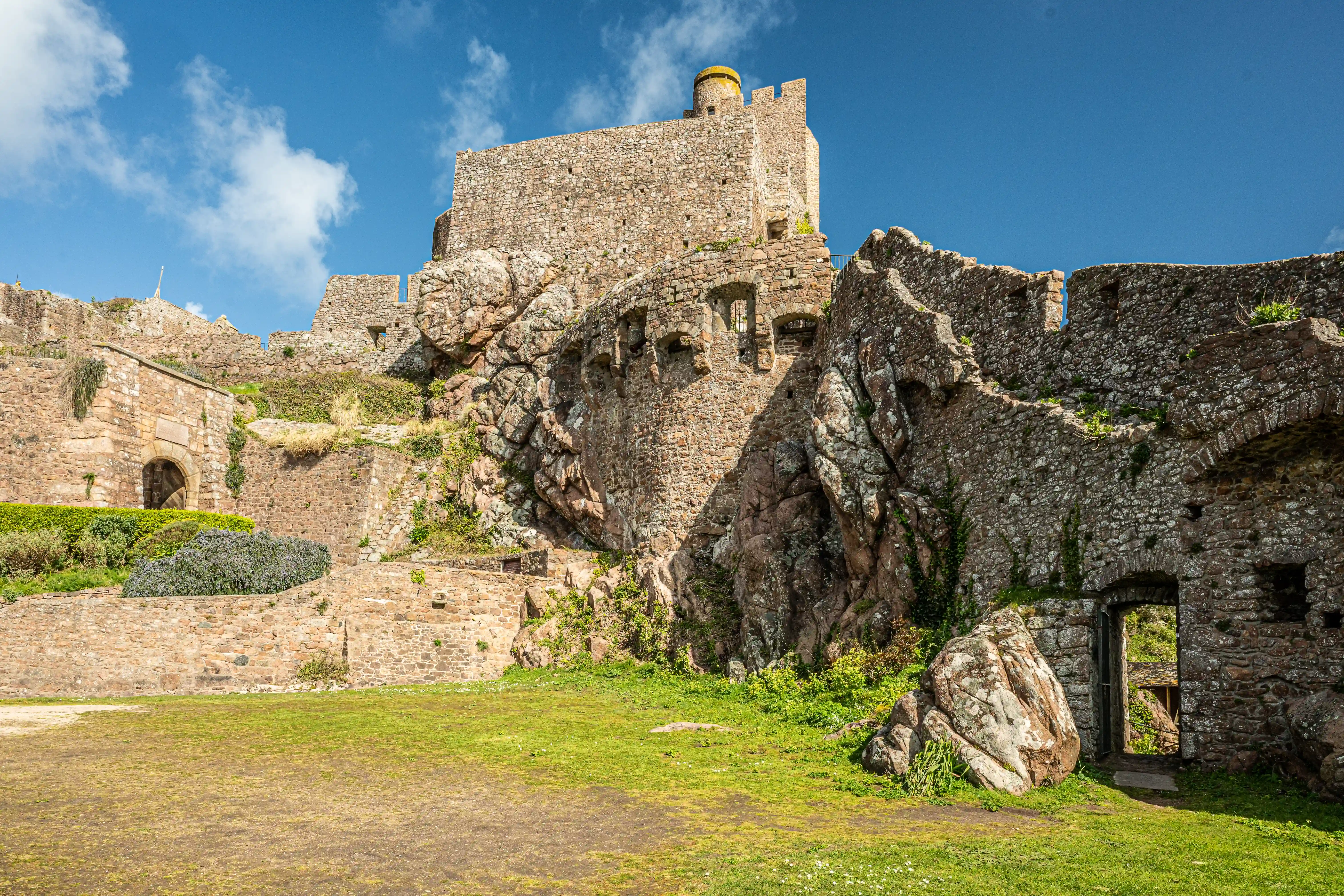 A wide view of a weathered stone castle ruin, built into a rocky hillside. The castle has stone walls, battlements, and arched doorways. The foreground is a grassy area, with a bright blue sky with white clouds above.