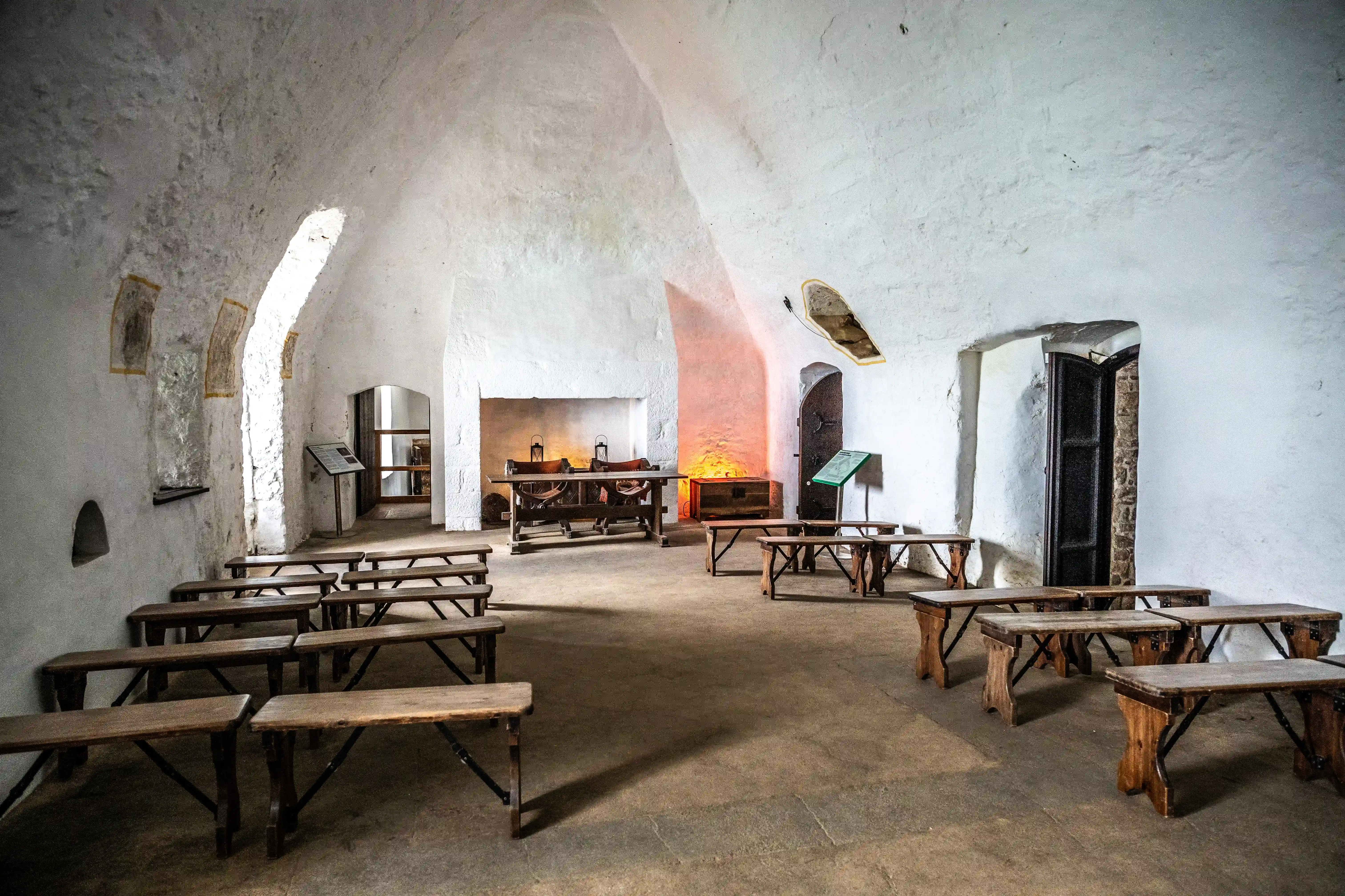 Interior of a historic room with white, textured walls, a vaulted ceiling, and rows of wooden benches. A large table is positioned in front of a fireplace, with warm light illuminating the background.