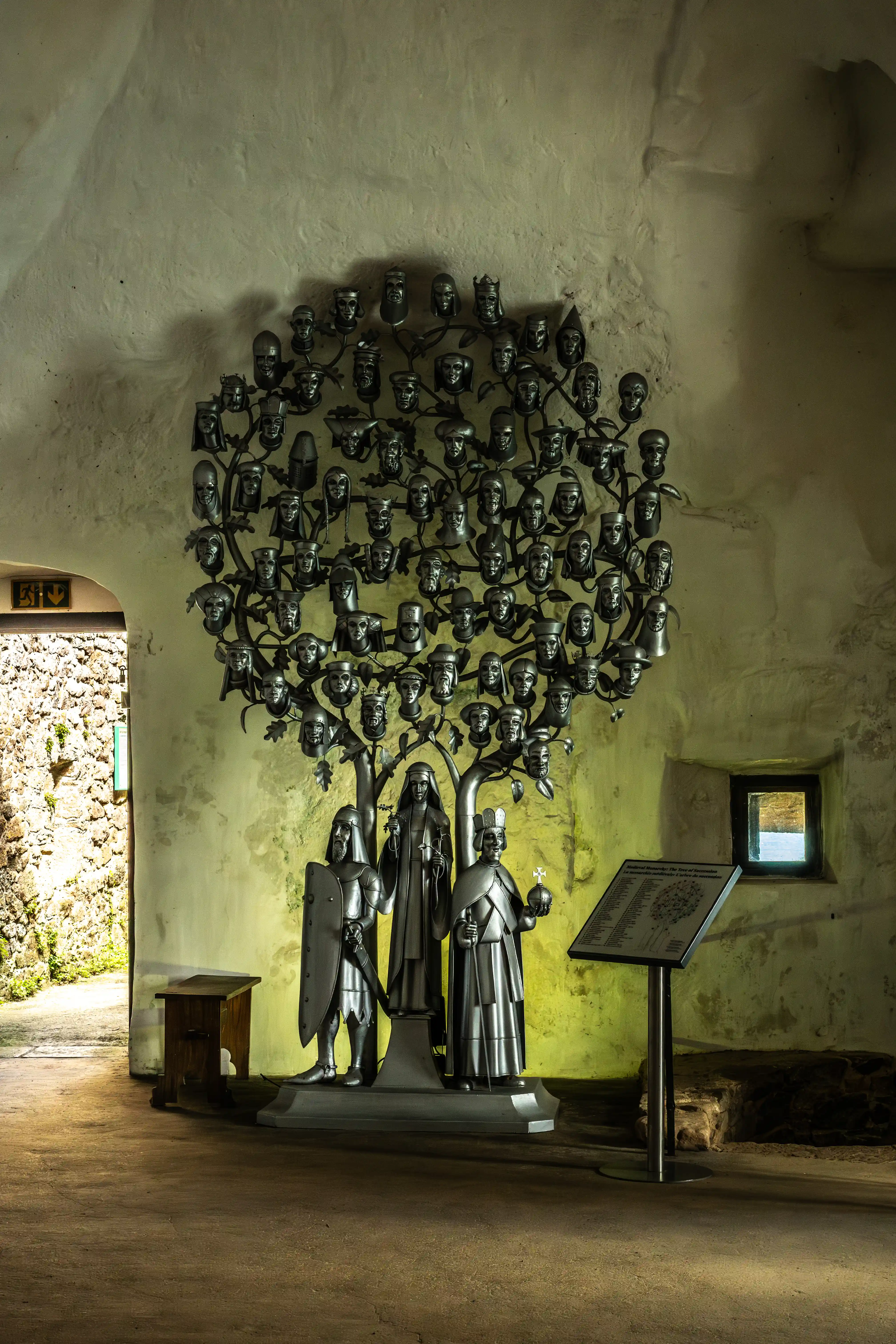 A metallic tree sculpture filled with many human heads, positioned centrally. To its left stands a knight with a shield, and to its right are two figures in medieval robes. A sign with text and a diagram stands to the right of the figures. The artwork is in a stone building with a doorway leading outside.