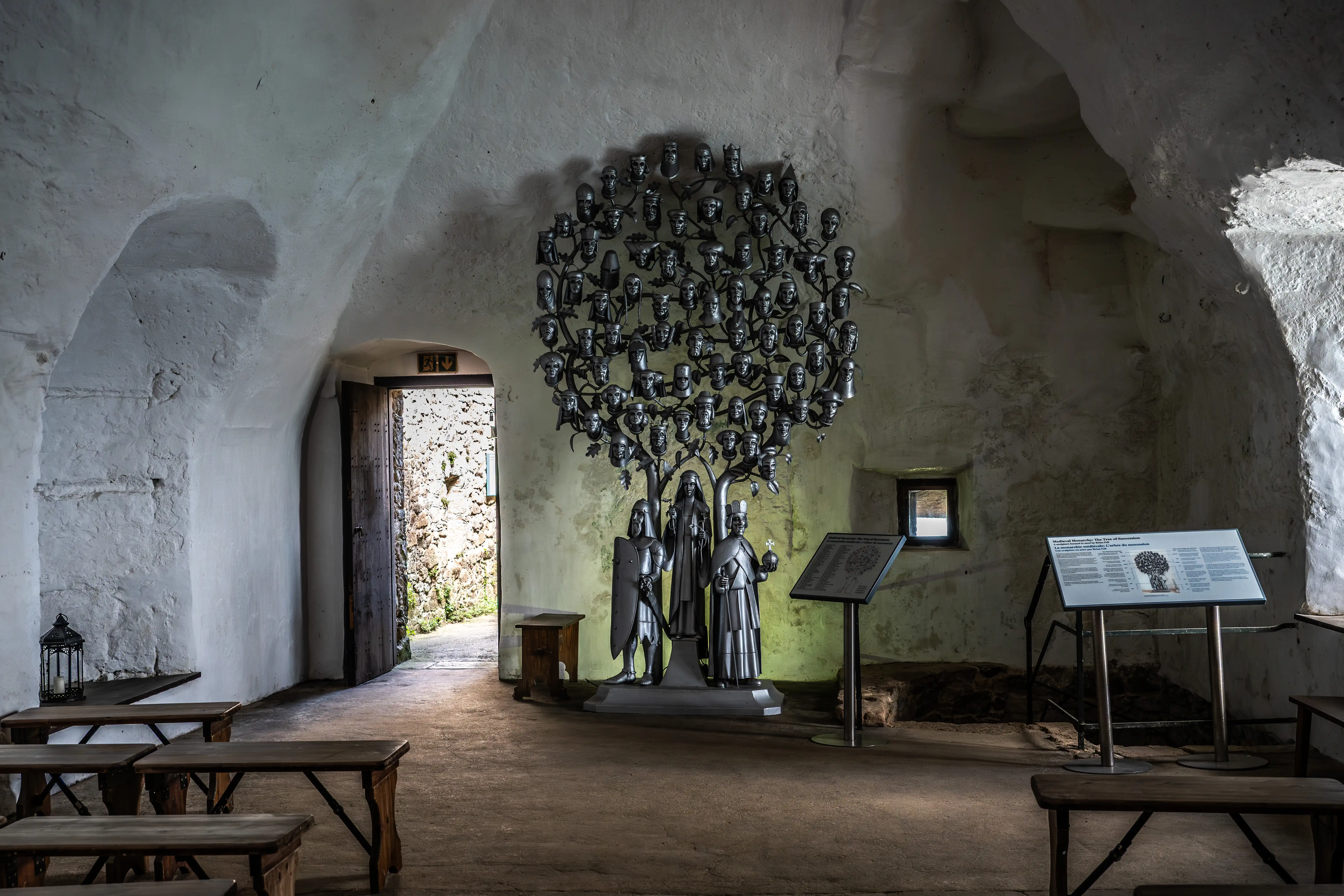  A metallic sculpture of a family tree with many heads as its leaves, stands in a room with rough stone walls. Three figures are at its base. Wooden benches and an exit door are visible.