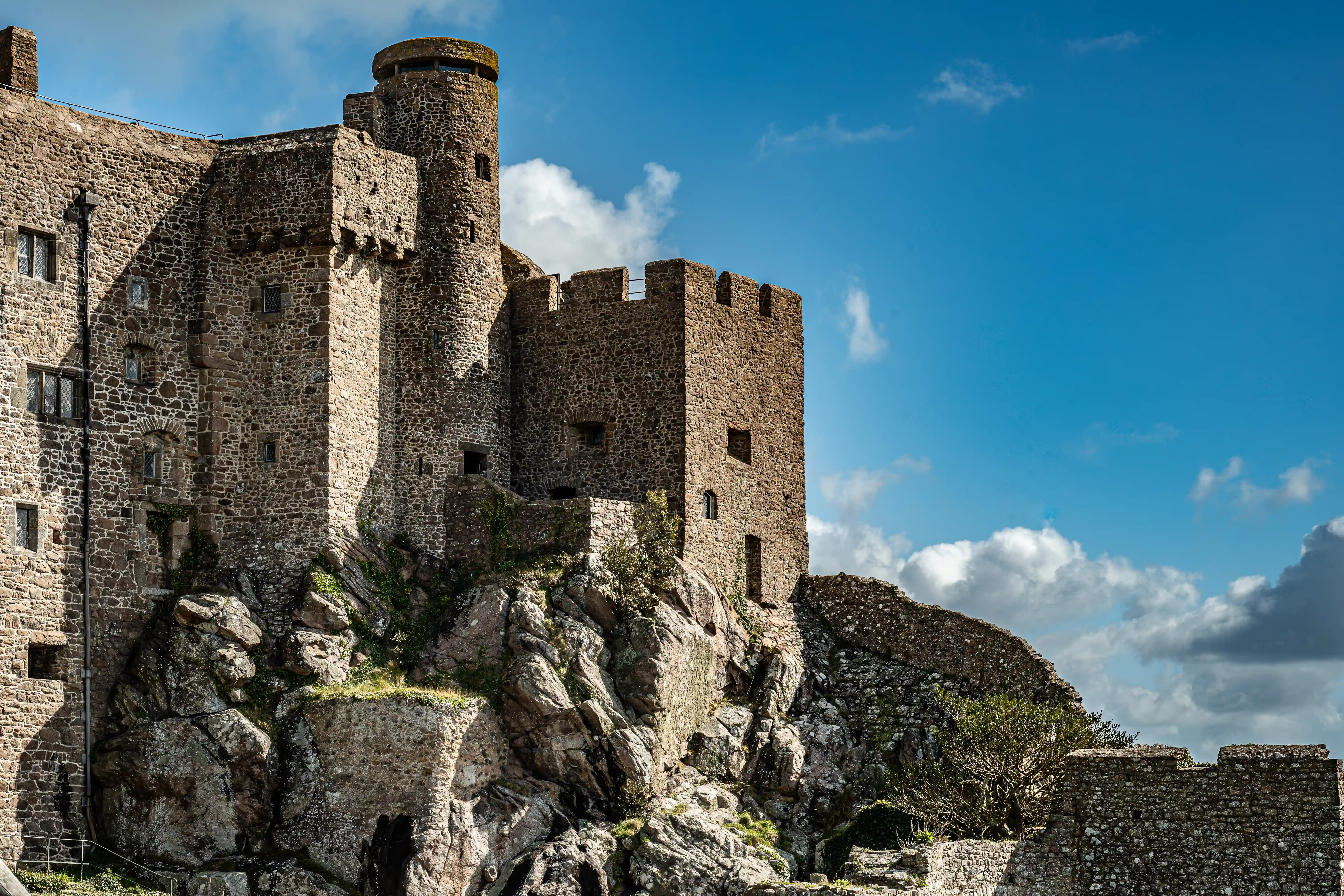A medieval stone castle with multiple towers and walls sits on a rugged rocky cliff. The sky above is a bright blue with scattered white clouds.
