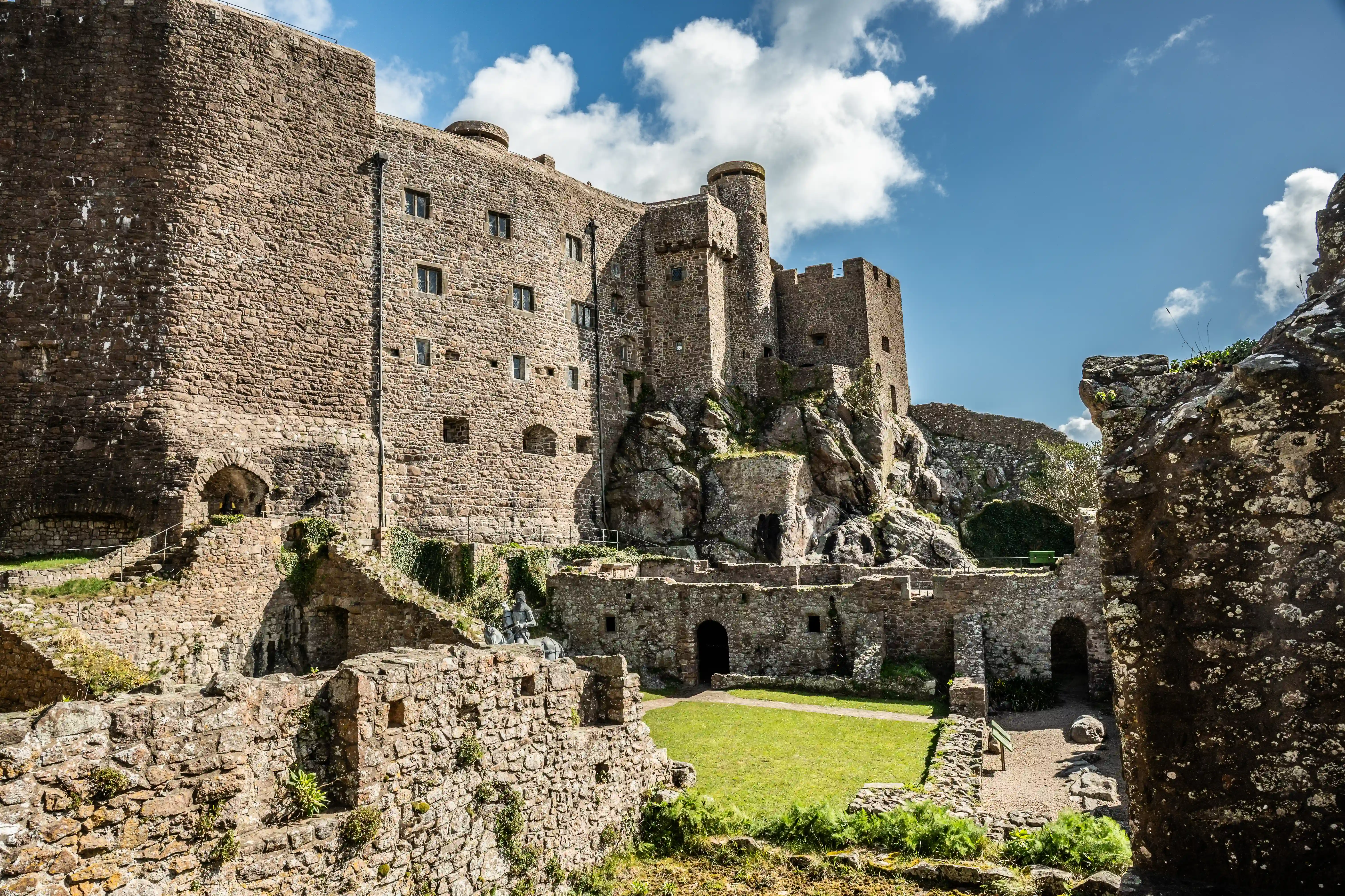 A panoramic view of a historic stone castle with multiple towers, built on a rocky hillside, featuring a green courtyard and a bright blue sky with clouds.