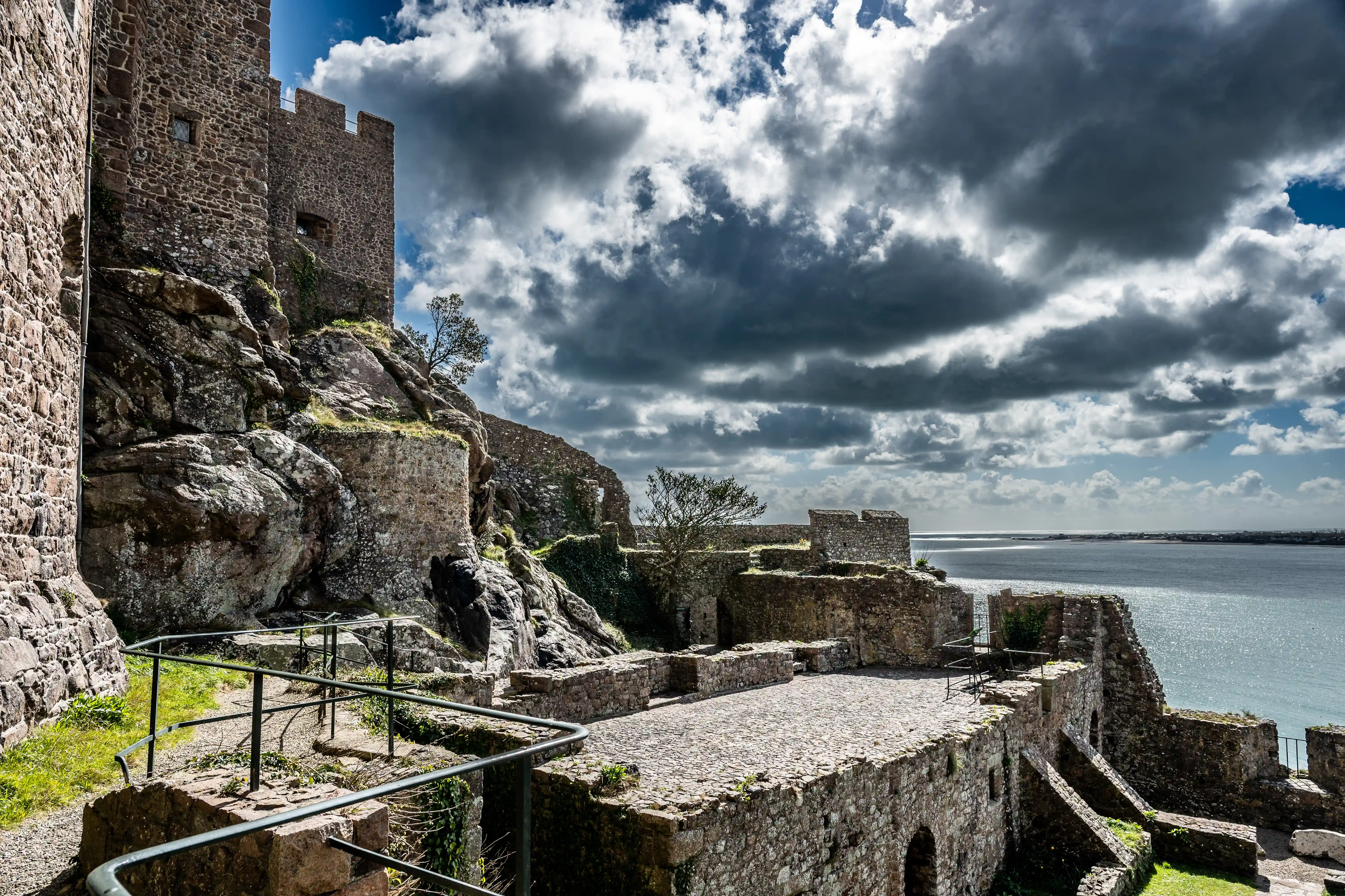 Stone castle ruins on a rocky outcrop beside a shimmering sea under a sky of dark, dramatic clouds and bright blue patches.