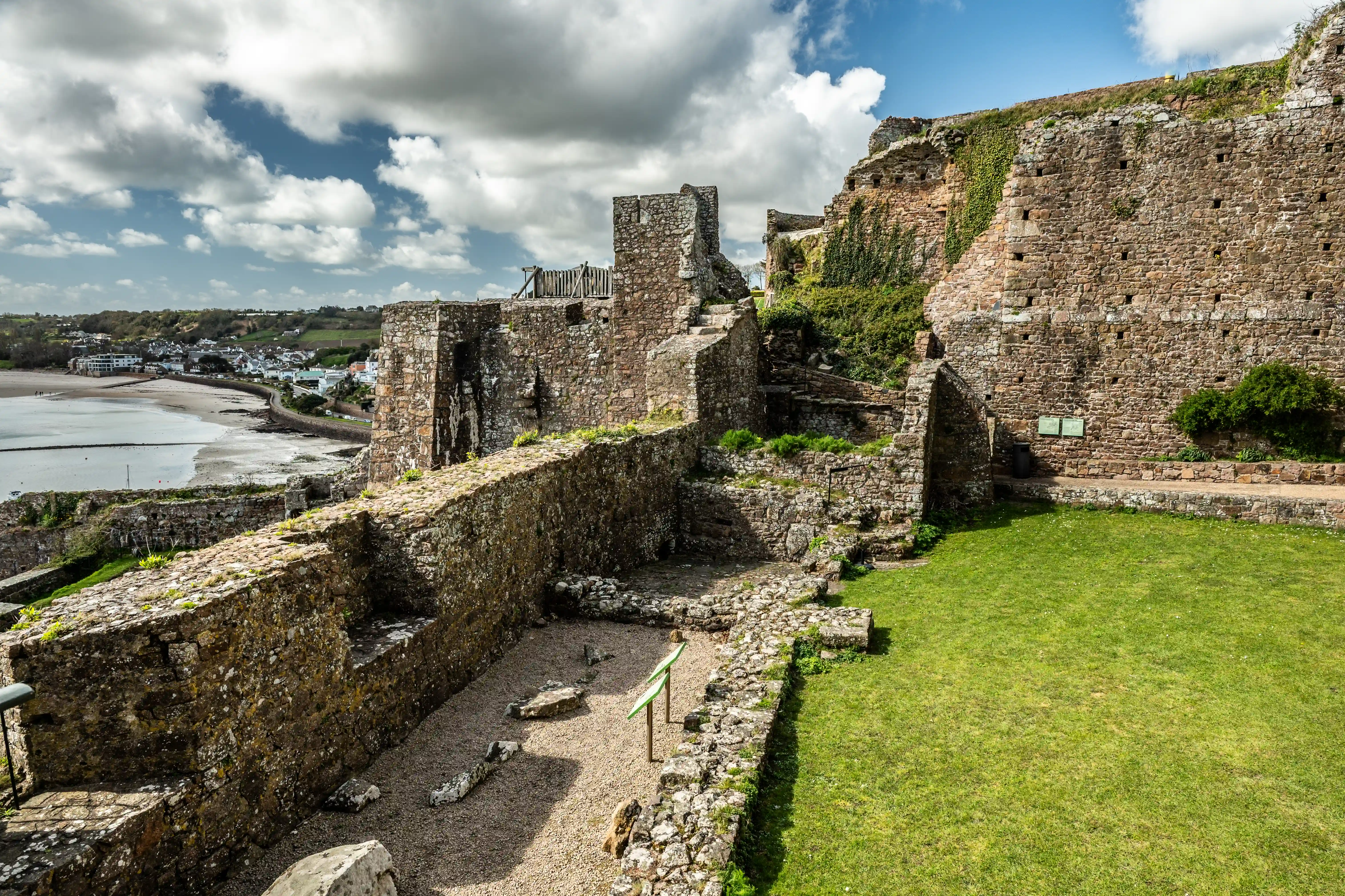 Ancient stone castle ruins with weathered walls and crumbling towers, overlooking a coastal village and bay under a bright blue sky with dramatic clouds.