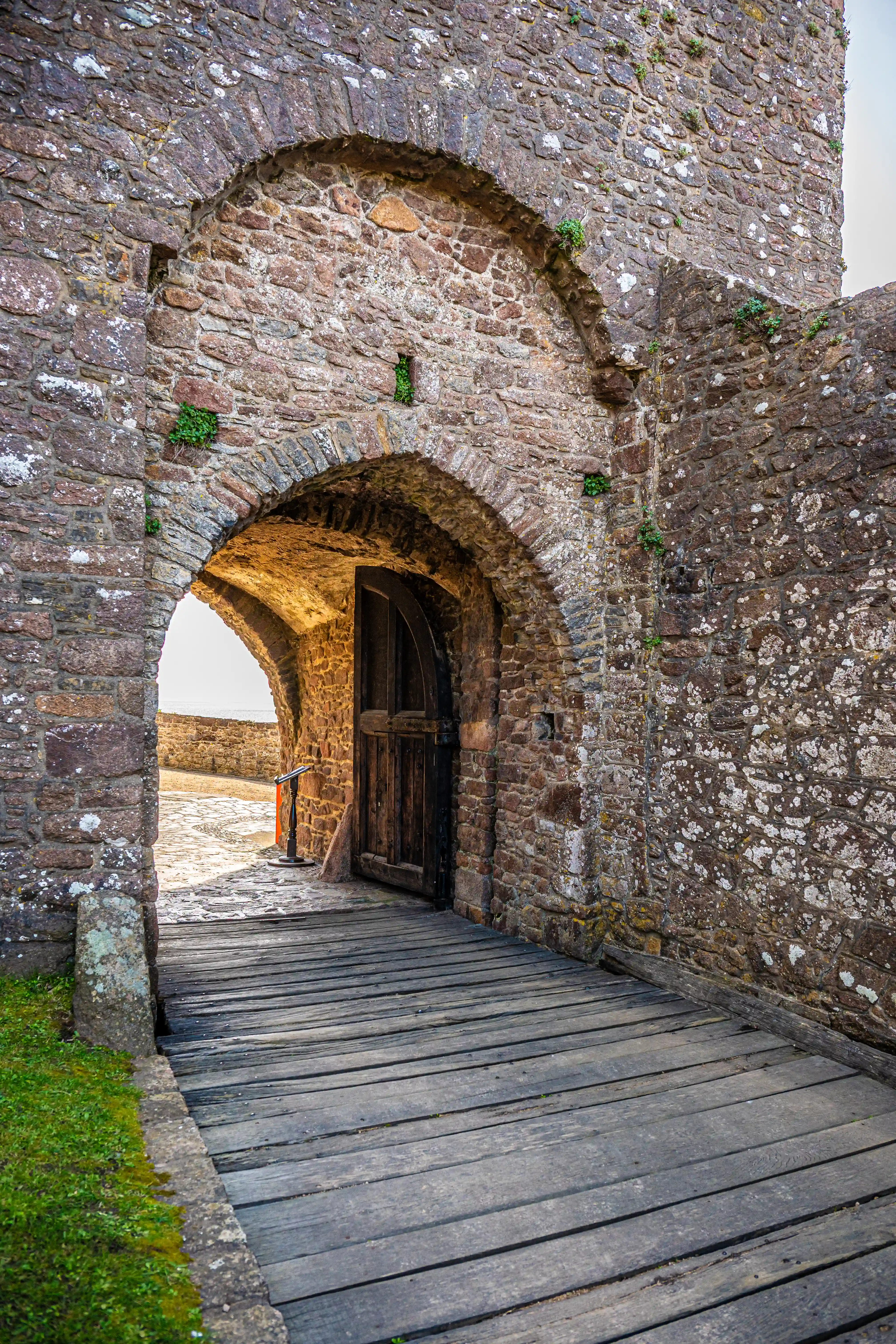A sturdy, arched stone gateway of an old castle, with a wooden drawbridge leading up to a large, dark wooden door. Small green plants grow from crevices in the stone walls.