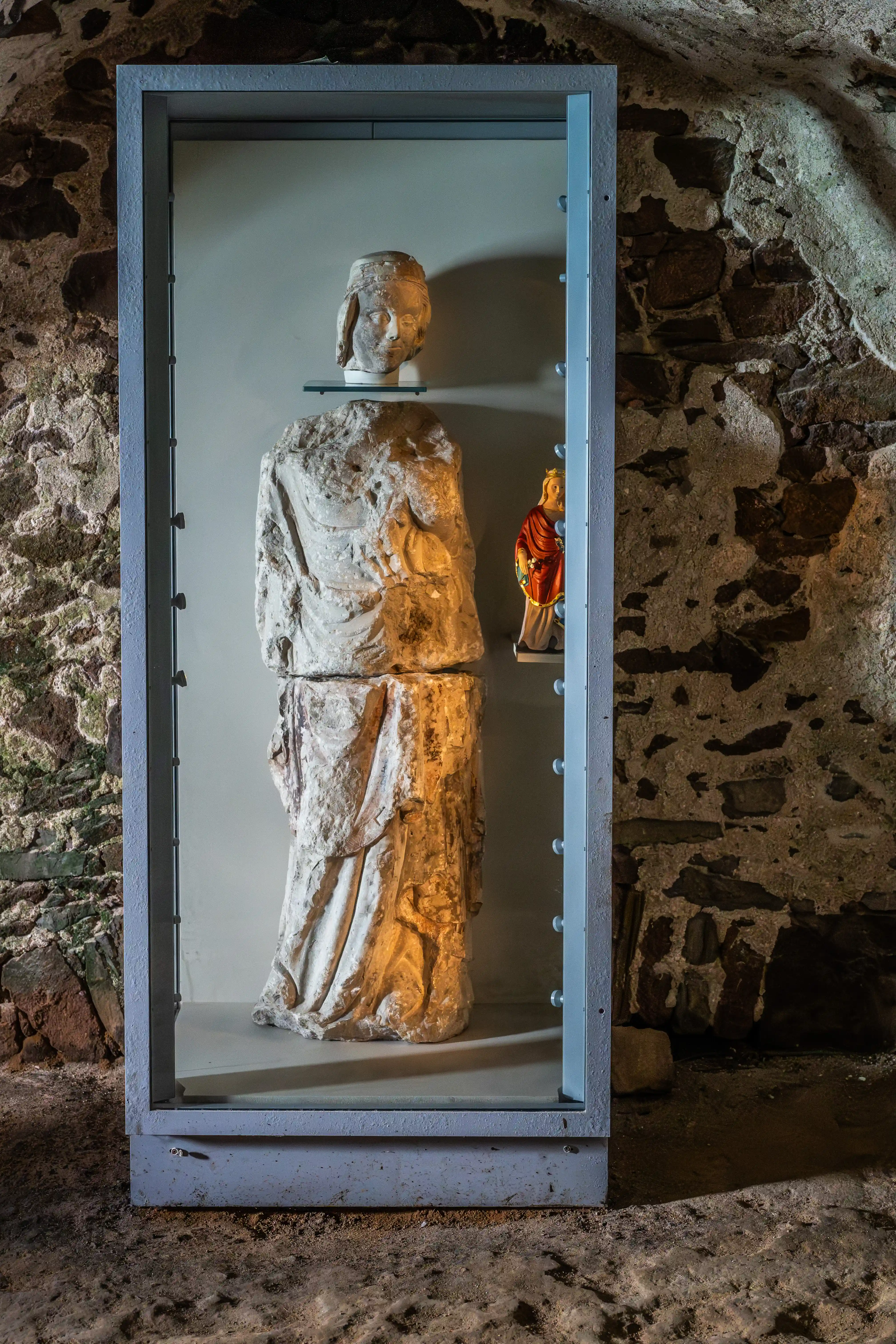 A broken ancient stone statue of a figure, with its head placed above the torso, is exhibited in a glass case. A small, colourful figurine is also partially visible. The display is set against a backdrop of ancient stone walls.