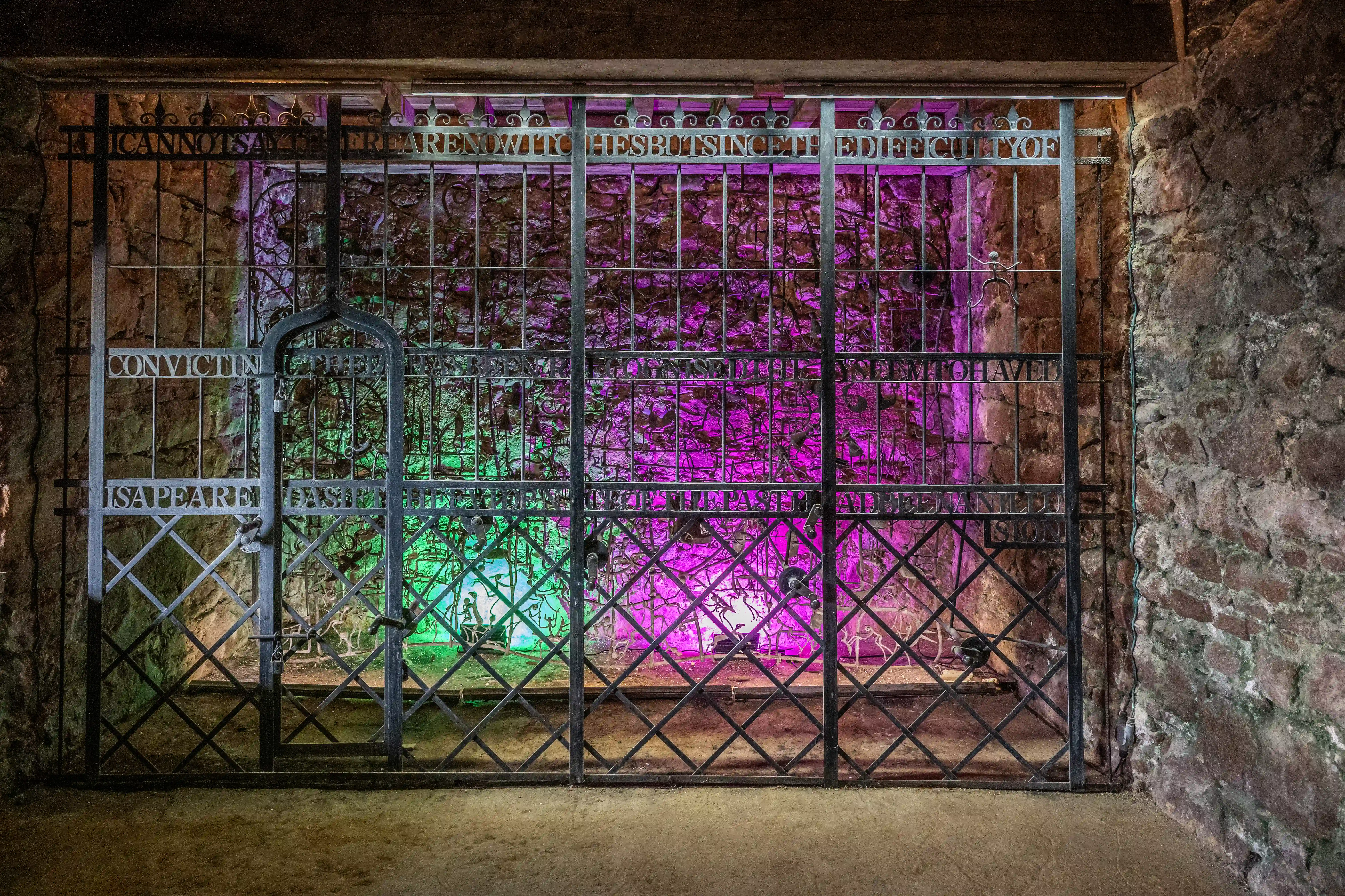 A dark, textured stone passageway leads to a striking wrought iron gate, intricately decorated with swirling patterns and small human-like figures.