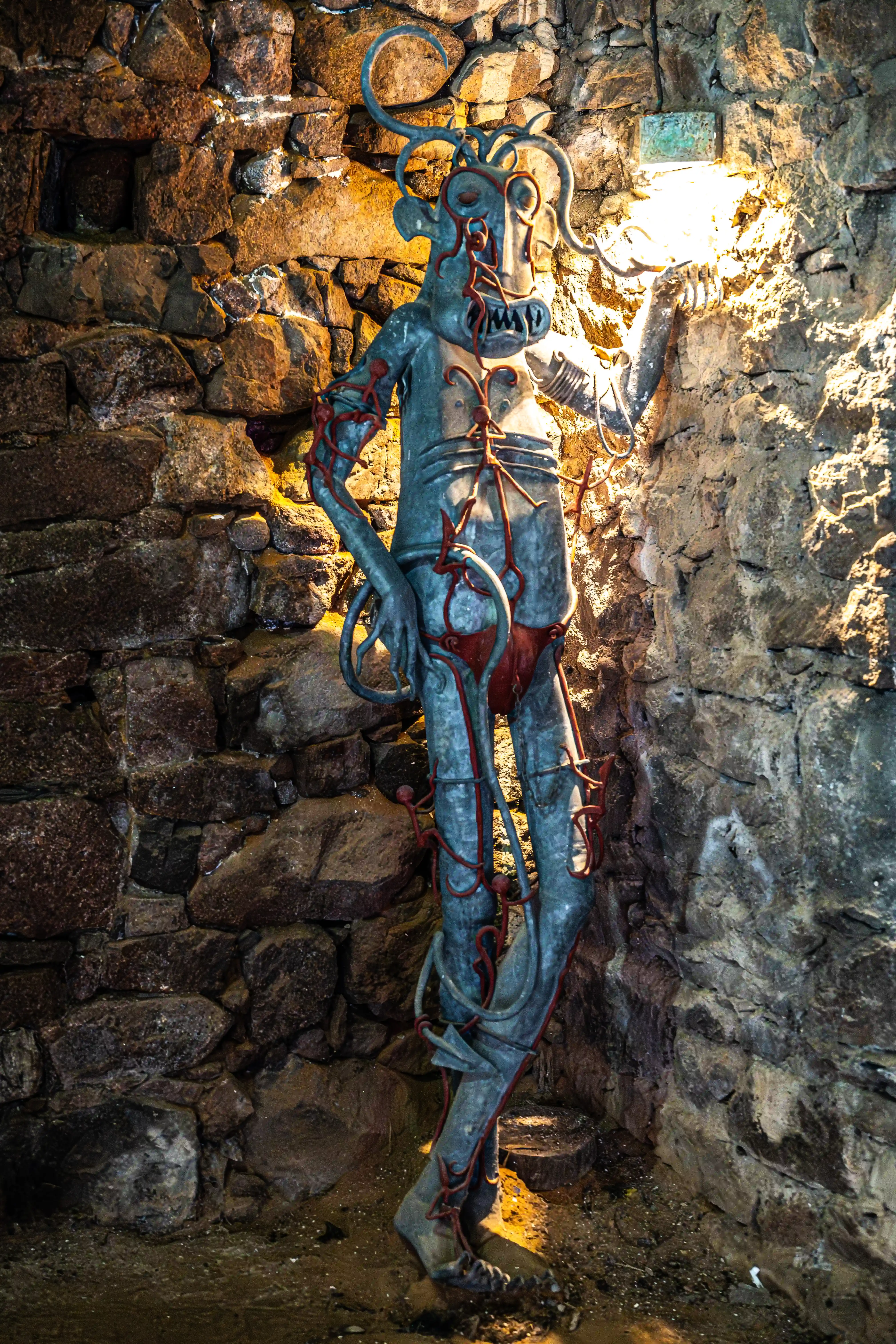 A tall, thin, metal sculpture of a grotesque figure with a grimacing mouth and red tendril-like details, standing in a dimly lit grotto made of rough stone. A bright light illuminates the figure and the wall behind it.