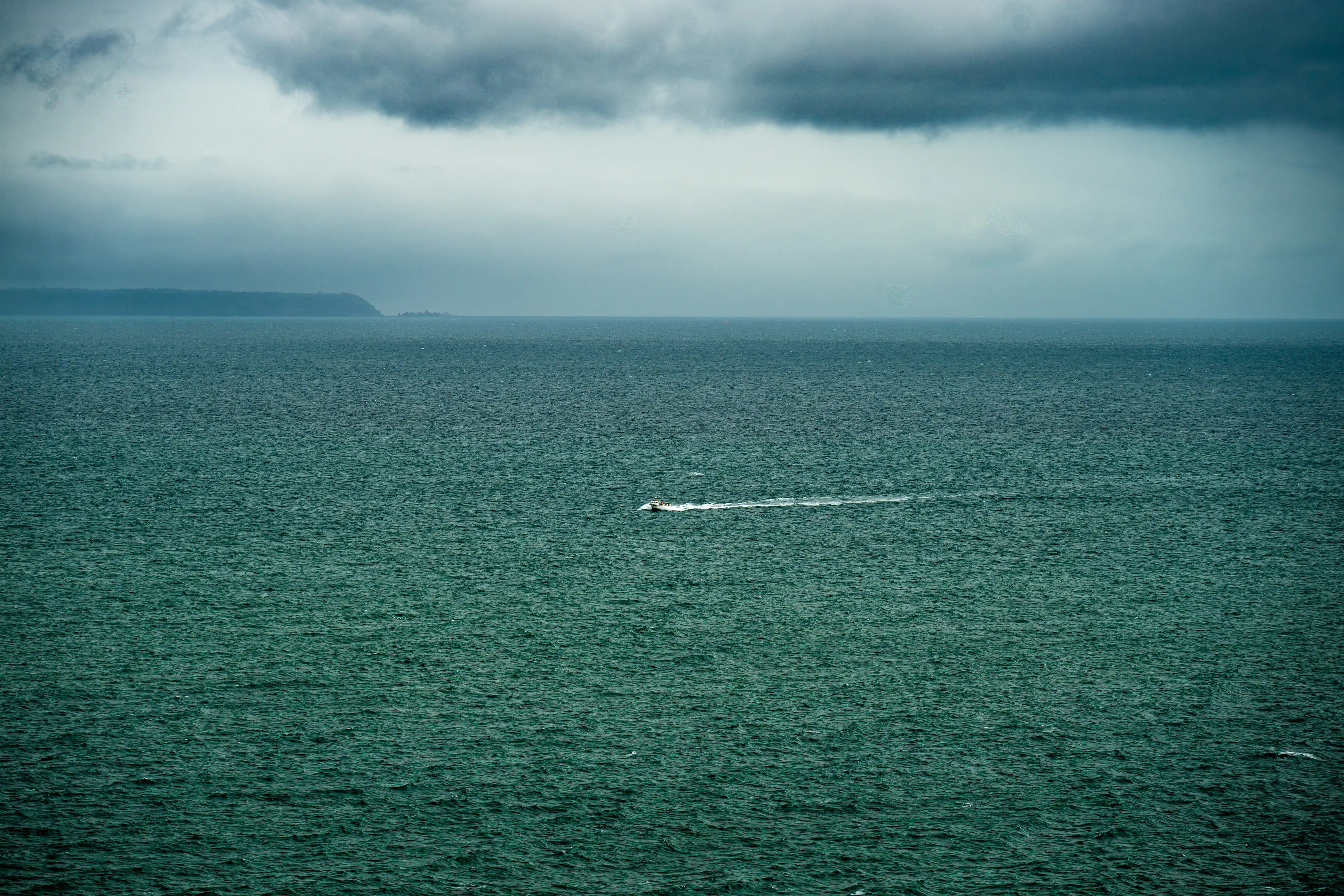 A small motorboat speeds across a choppy, dark teal ocean. In the distance, a faint, dark line of land is visible under a heavy, overcast sky with dramatic clouds.