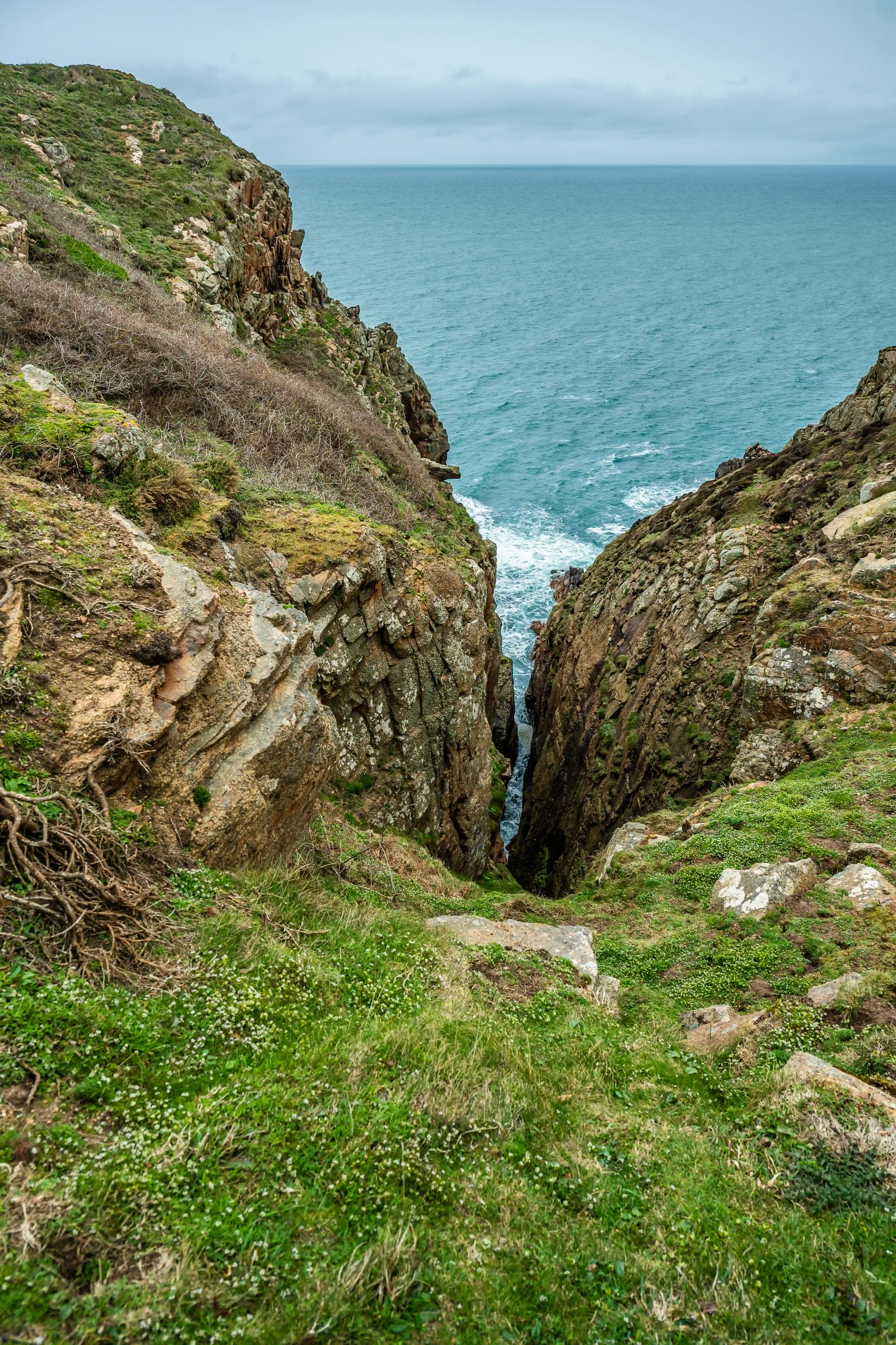 A vertical, wide-angle shot looking down a steep, narrow coastal gorge. Rocky cliffs covered in patches of green grass and moss flank the passage. The sea, a deep blue with white-capped waves, is visible at the bottom, beyond the end of the gorge. The sky above is a pale, cloudy grey