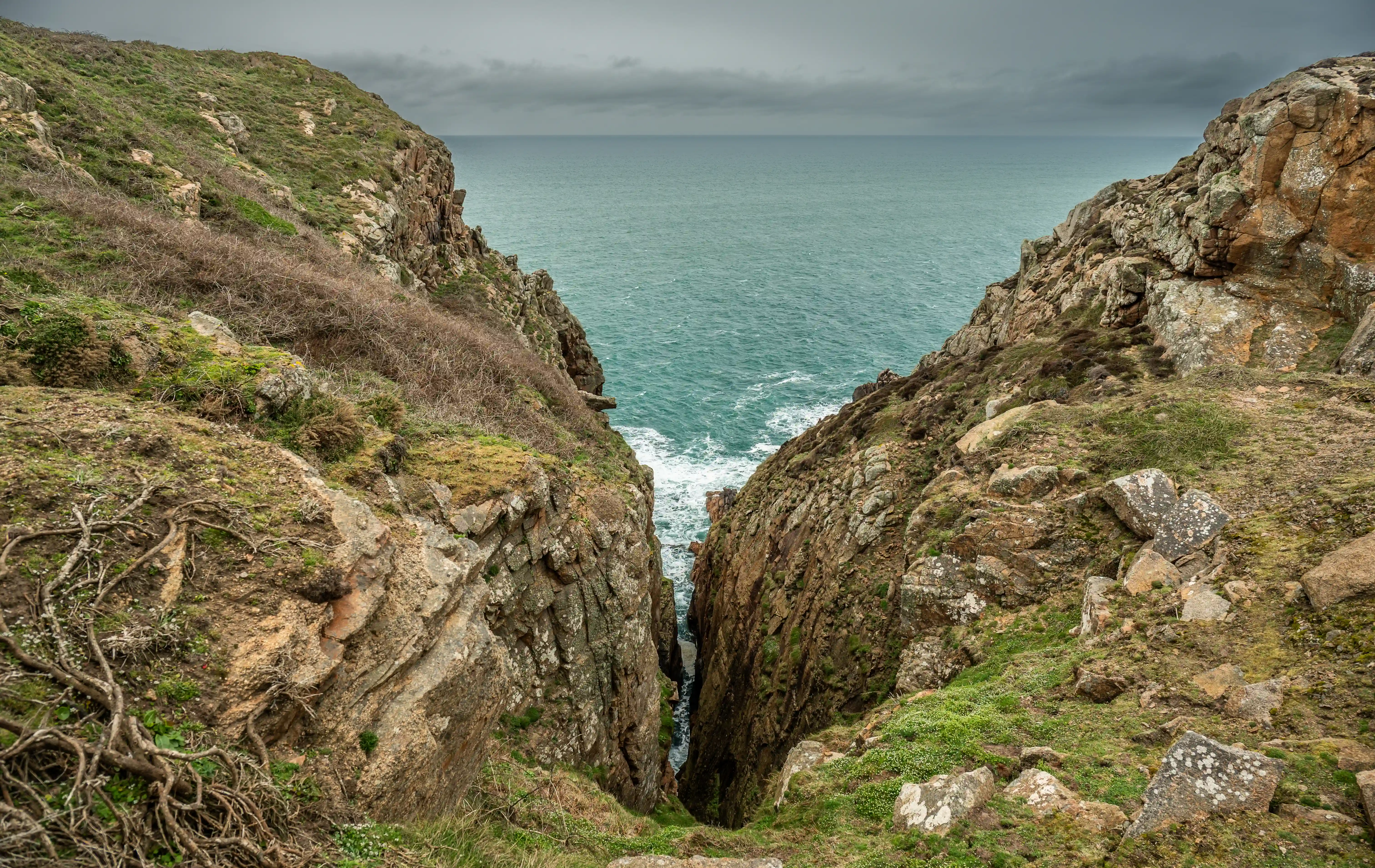 A steep, rocky coastal cliff face is split by a deep, dark chasm leading to the sea. The sea is a vibrant turquoise colour with white waves crashing at the bottom of the chasm and further out. The sky is overcast and grey.