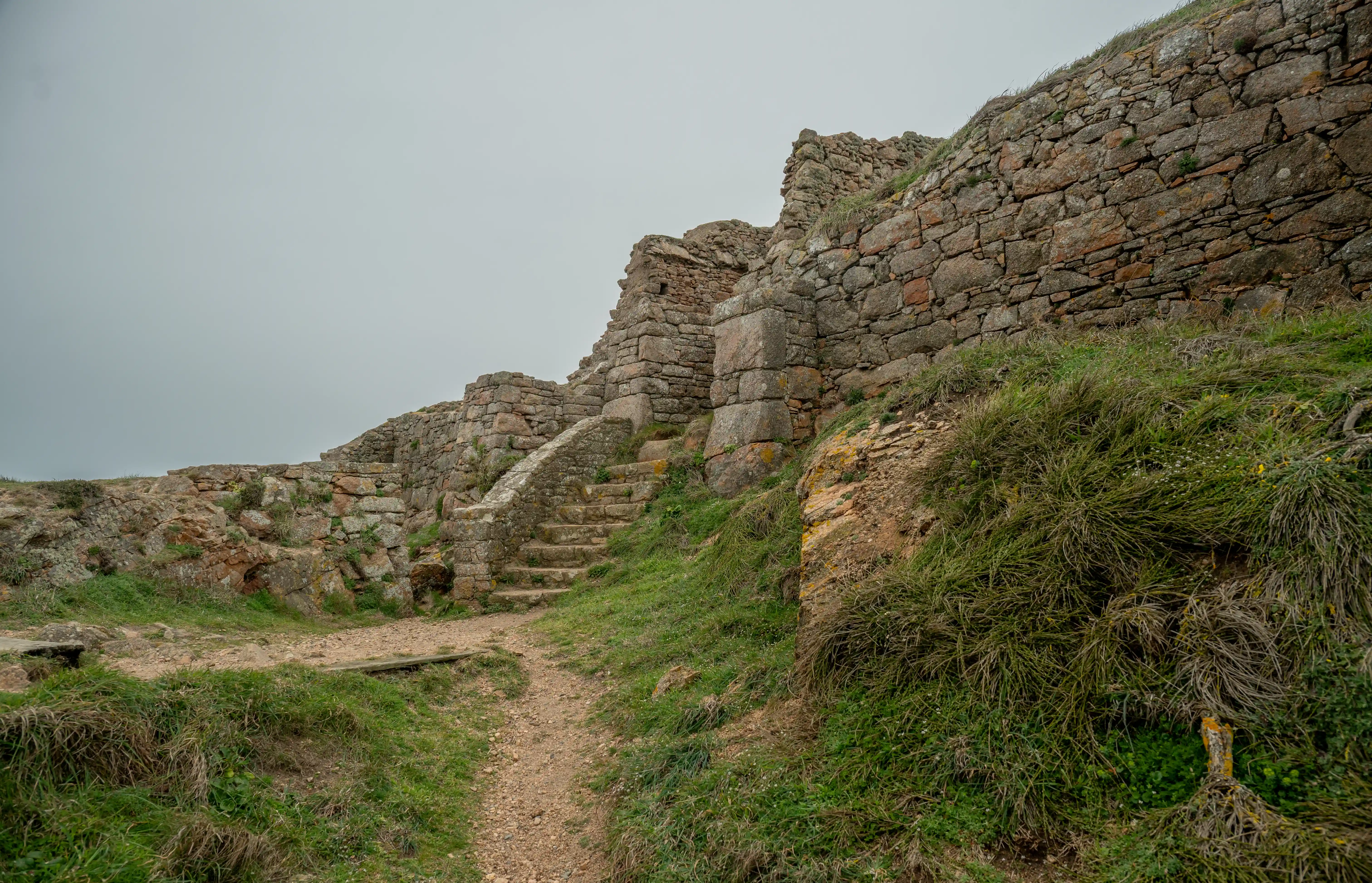 Stone ruins of a wall and staircase on a grassy, rocky hill under a grey sky.