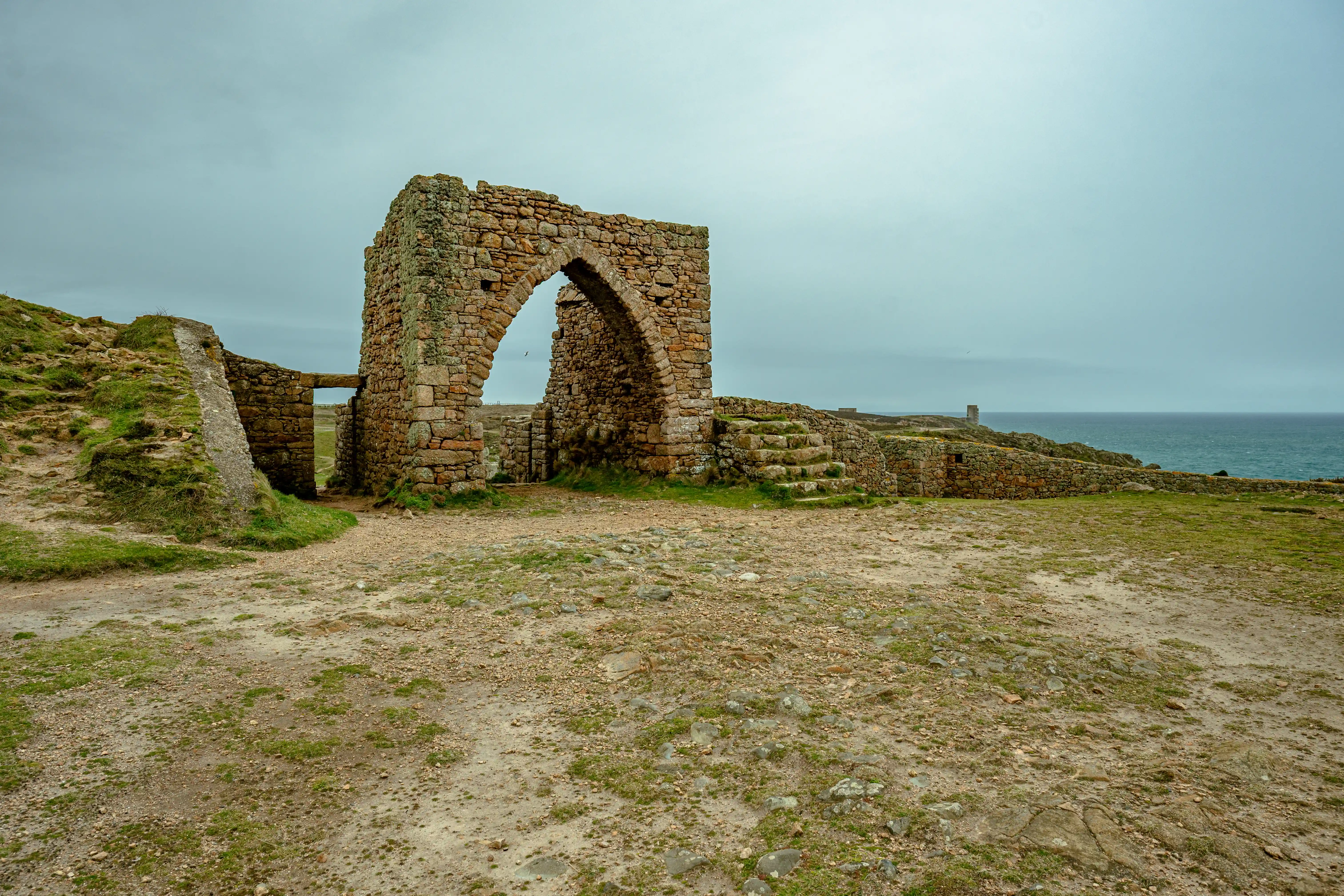 Stone archway ruins on a coastal headland, with grassy ground and the sea in the background under a cloudy sky.