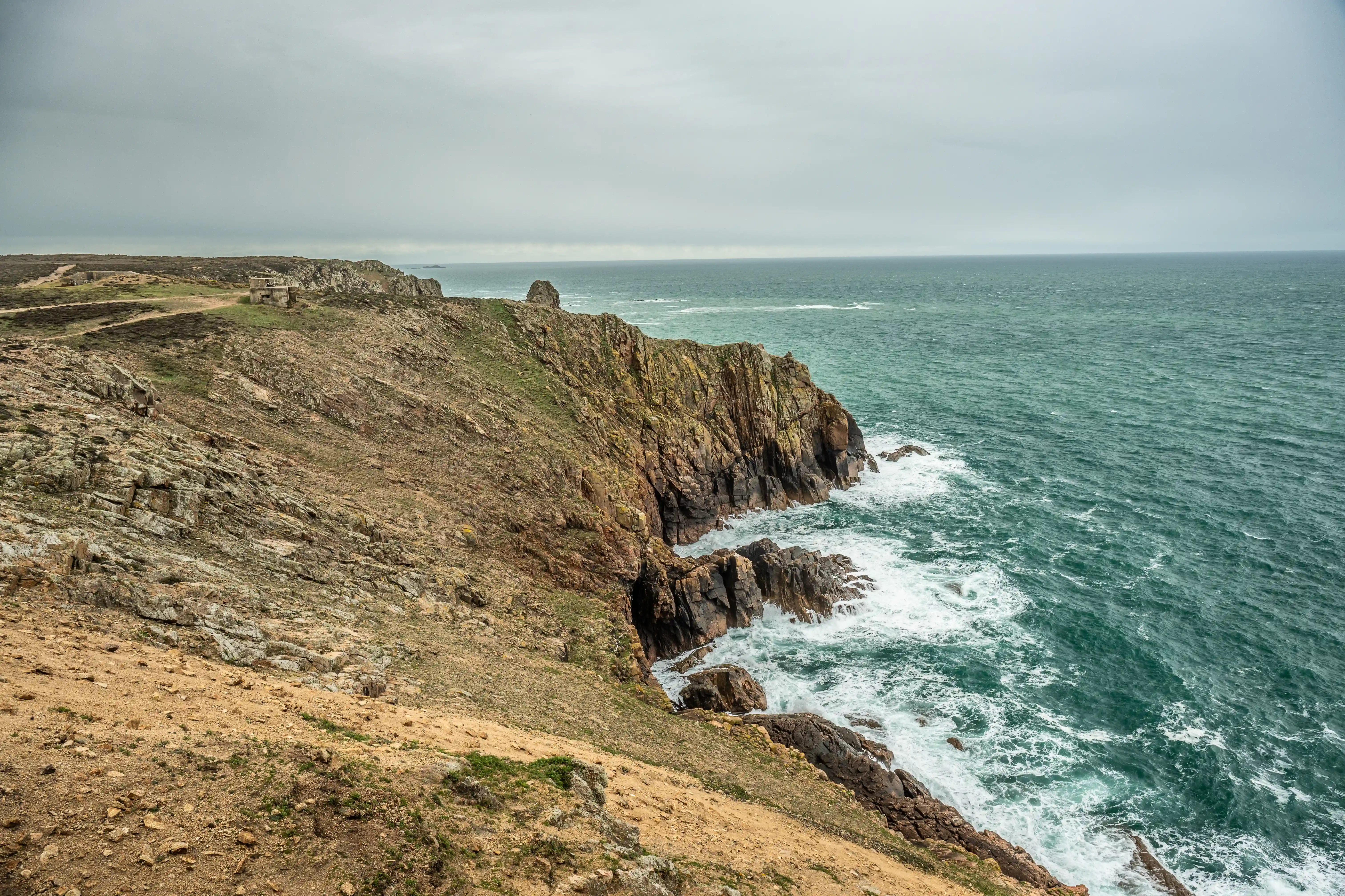 A panoramic view of a rocky, green-topped coastline in Cornwall, England, with waves crashing against the cliffs. Several stone buildings are scattered on the plateau overlooking the sea, and the sky is overcast.