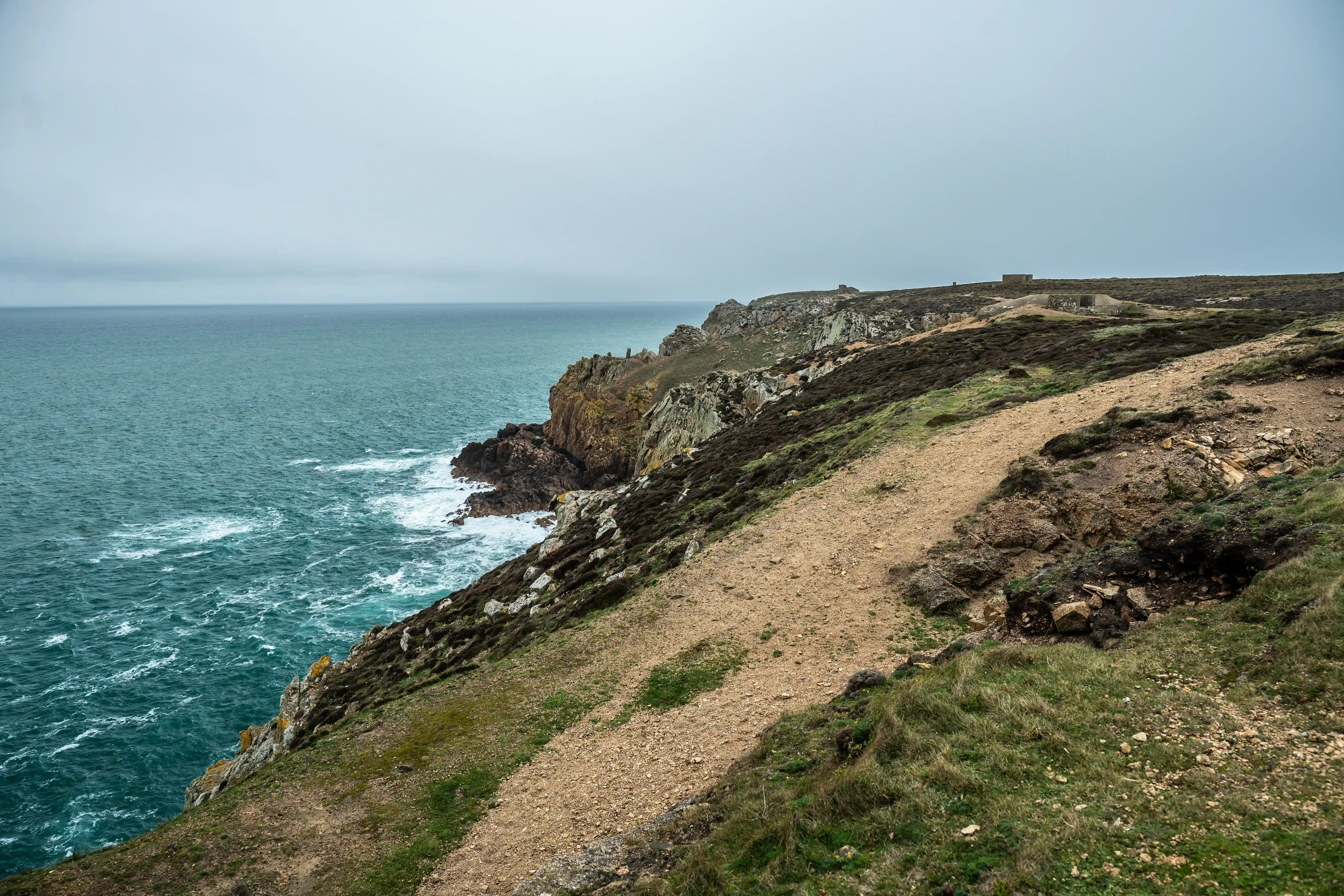 A dirt and grass path curves along a steep, rocky cliff face where the choppy, dark teal sea crashes against the shore, under a heavily overcast sky.