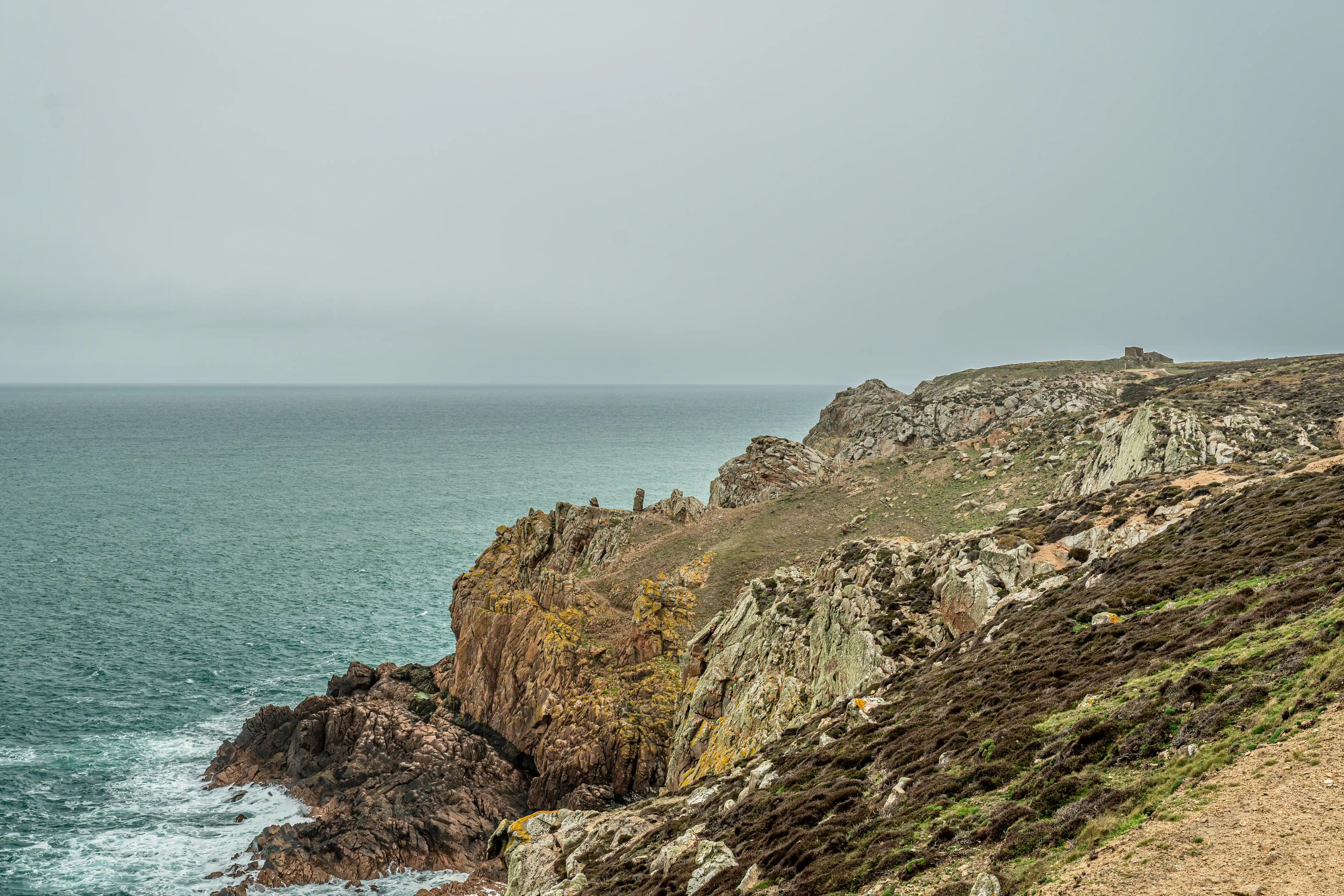 A panoramic view of a rocky coastline. The sea is to the left, with waves breaking against dark, jagged cliffs. The clifftop is covered in heathland and scattered rocks, with a small stone ruin visible on the far right horizon under a grey sky.