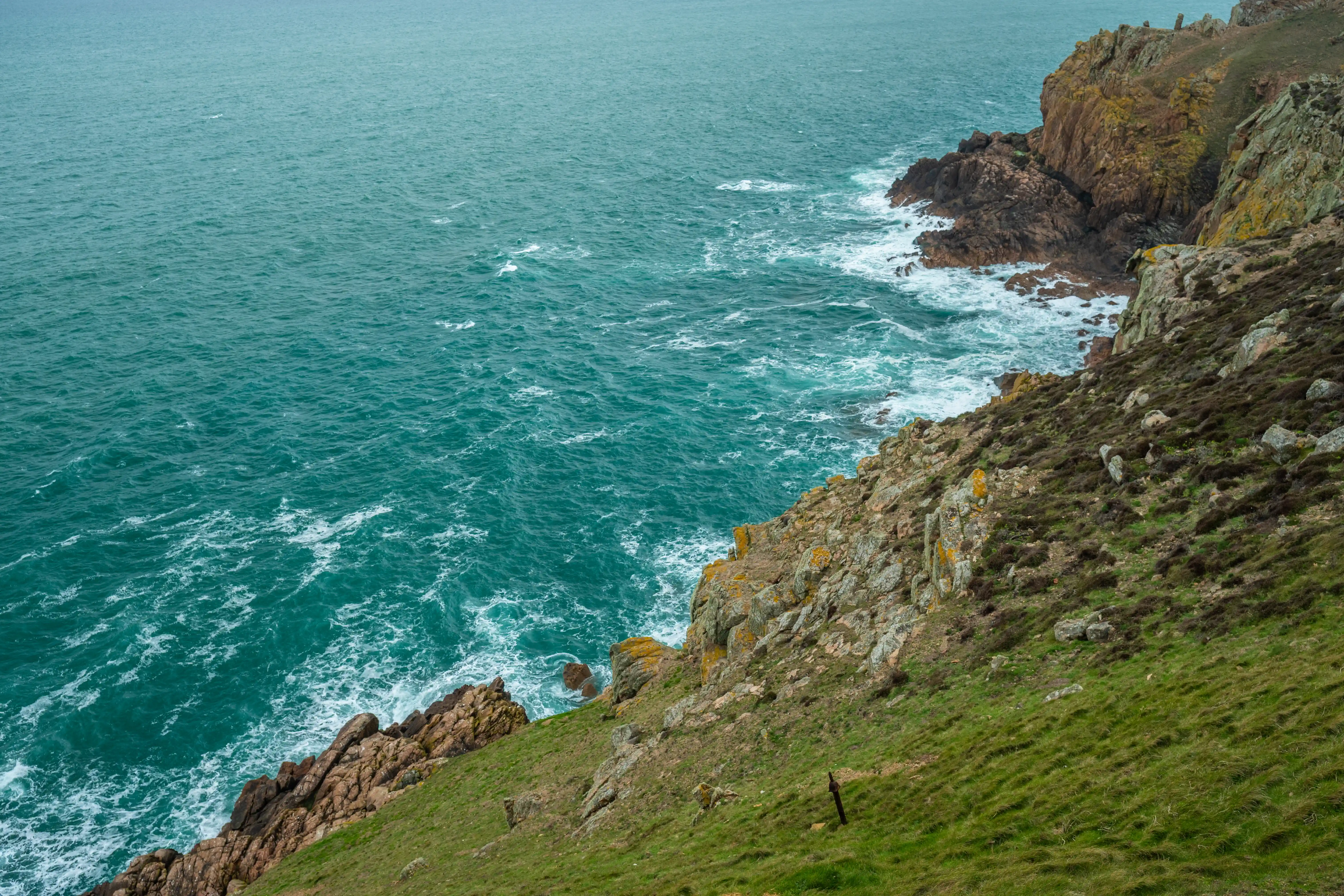 A high-angle view of a green, grassy cliff face sloping down towards a rough sea with white waves. Jagged, brown and grey rocks jut out into the water on the right side of the image.