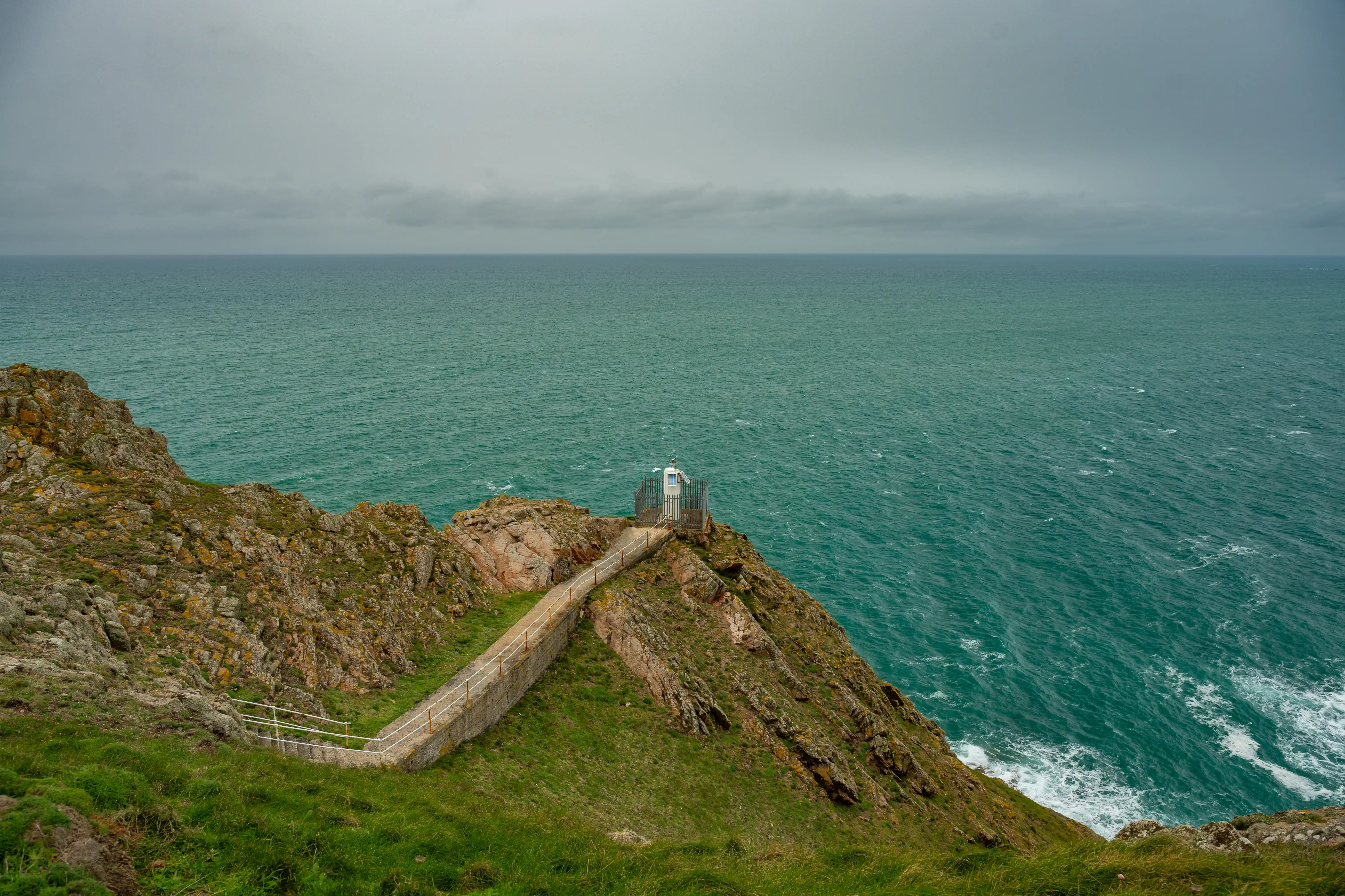 A winding coastal path leads to a small, modern observation structure on a rocky cliff overlooking a turbulent turquoise sea under a grey, cloudy sky.