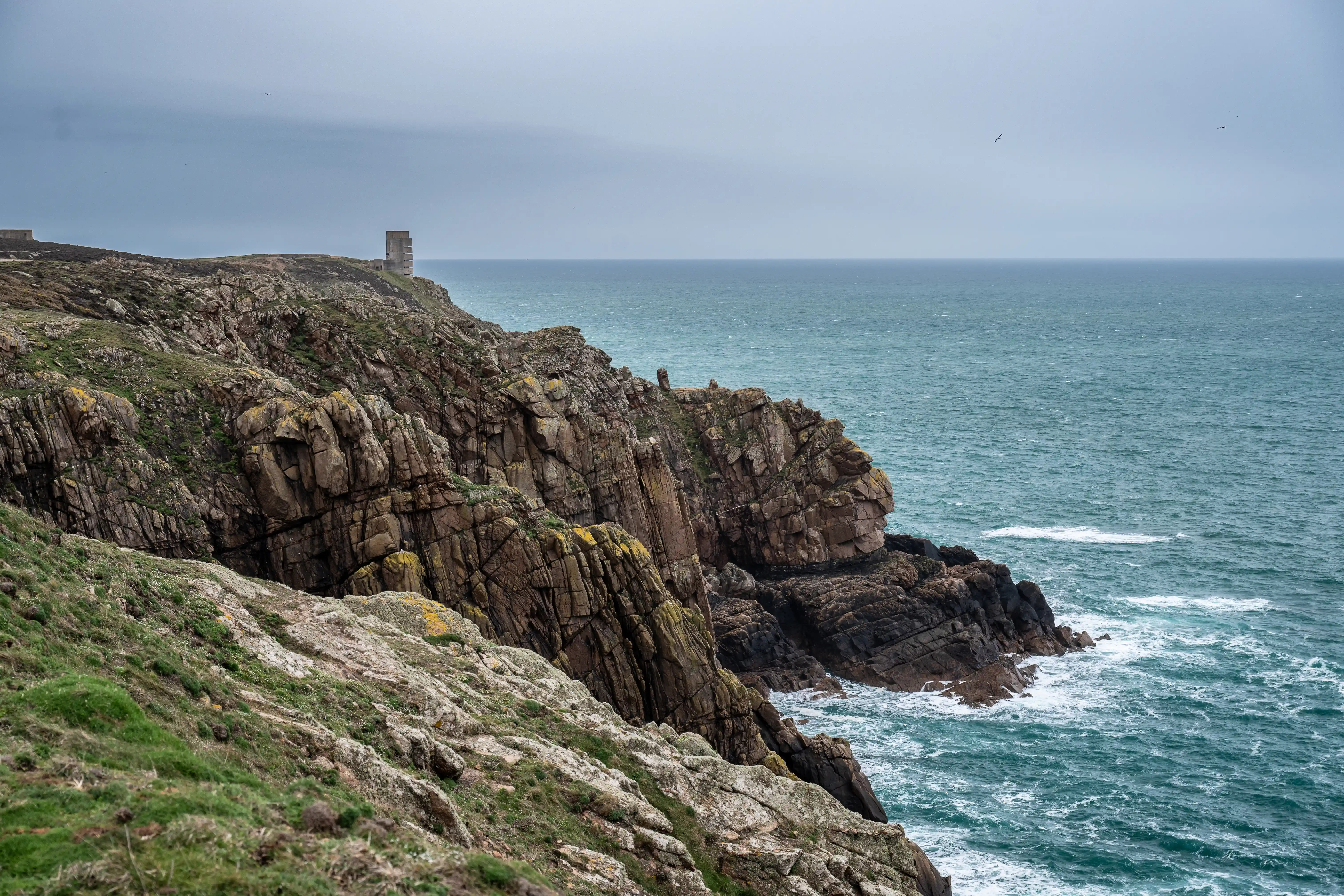 Wide shot of a dramatic, rocky coastline with steep cliffs and the turquoise ocean under a cloudy sky. A solitary old stone building is visible on the clifftop in the distance.