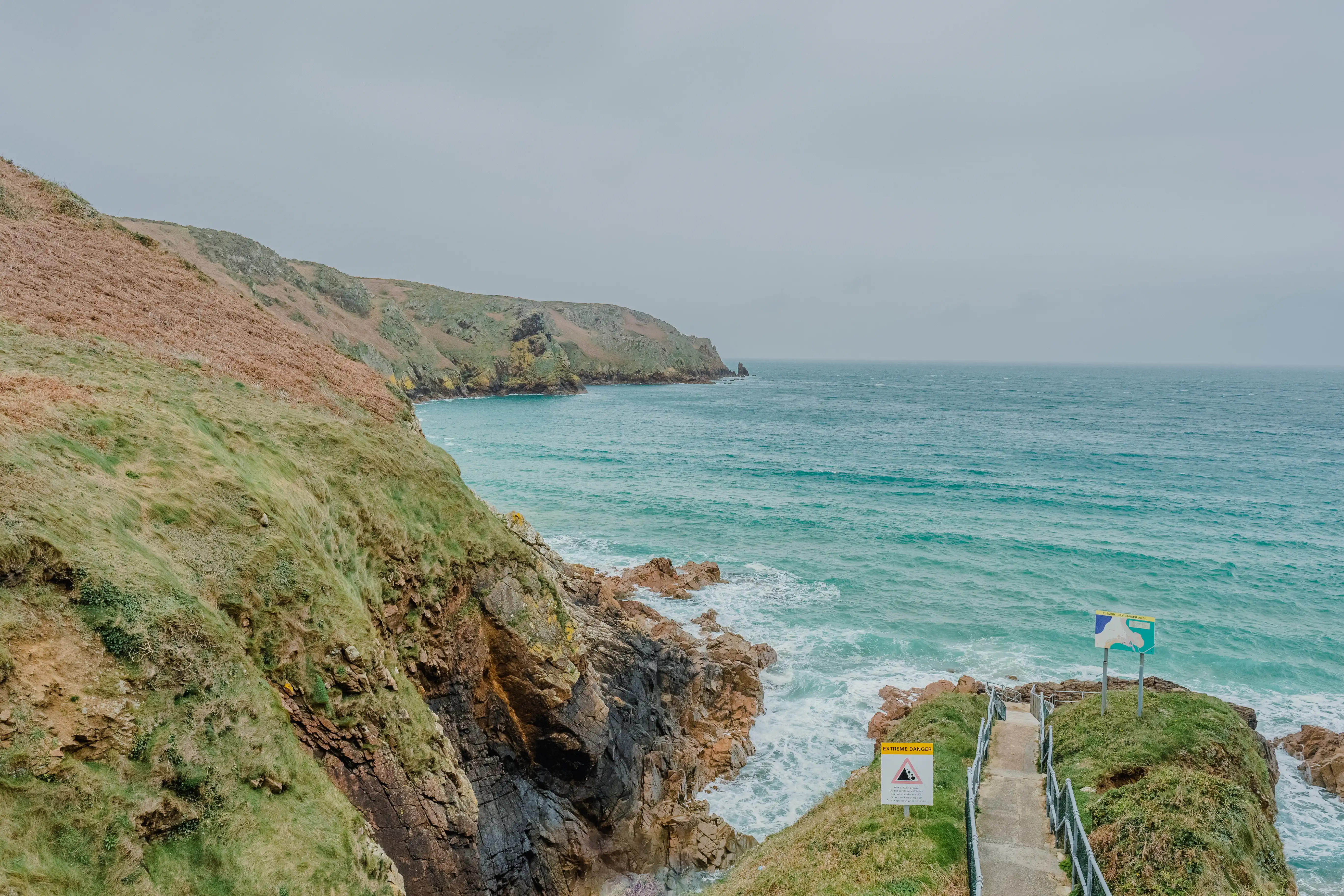 A pathway with metal railings leads over a grassy headland towards the sea, with steep, rocky cliffs on the left. Warning signs are visible along the path. The sea is a vibrant turquoise with white waves crashing against the rocks below.