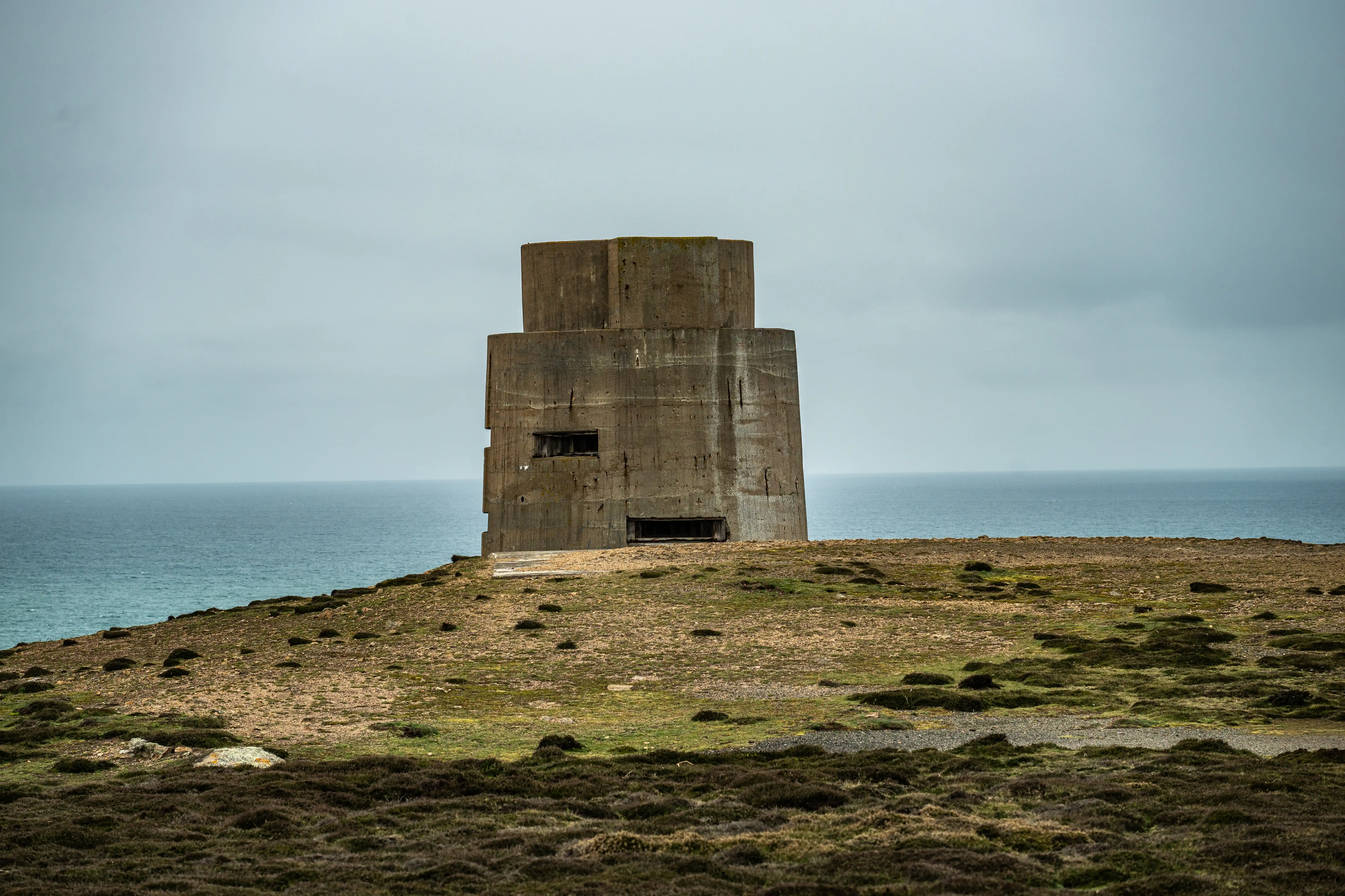 A weathered, multi-level concrete military defence tower situated on a grassy headland by the sea, with a cloudy sky overhead.