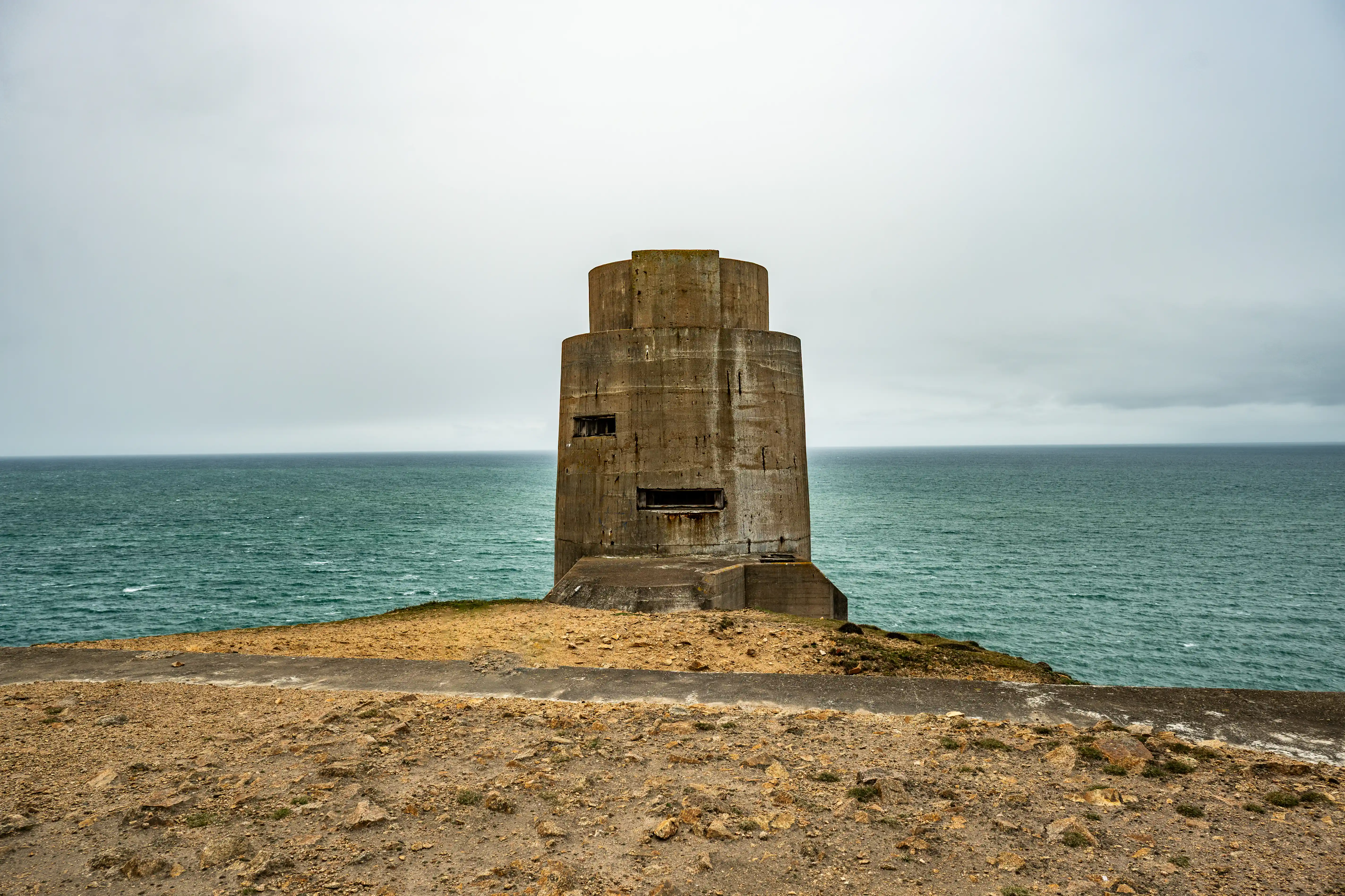 A weathered, multi-level concrete military defence tower situated on a grassy headland by the sea, with a cloudy sky overhead.