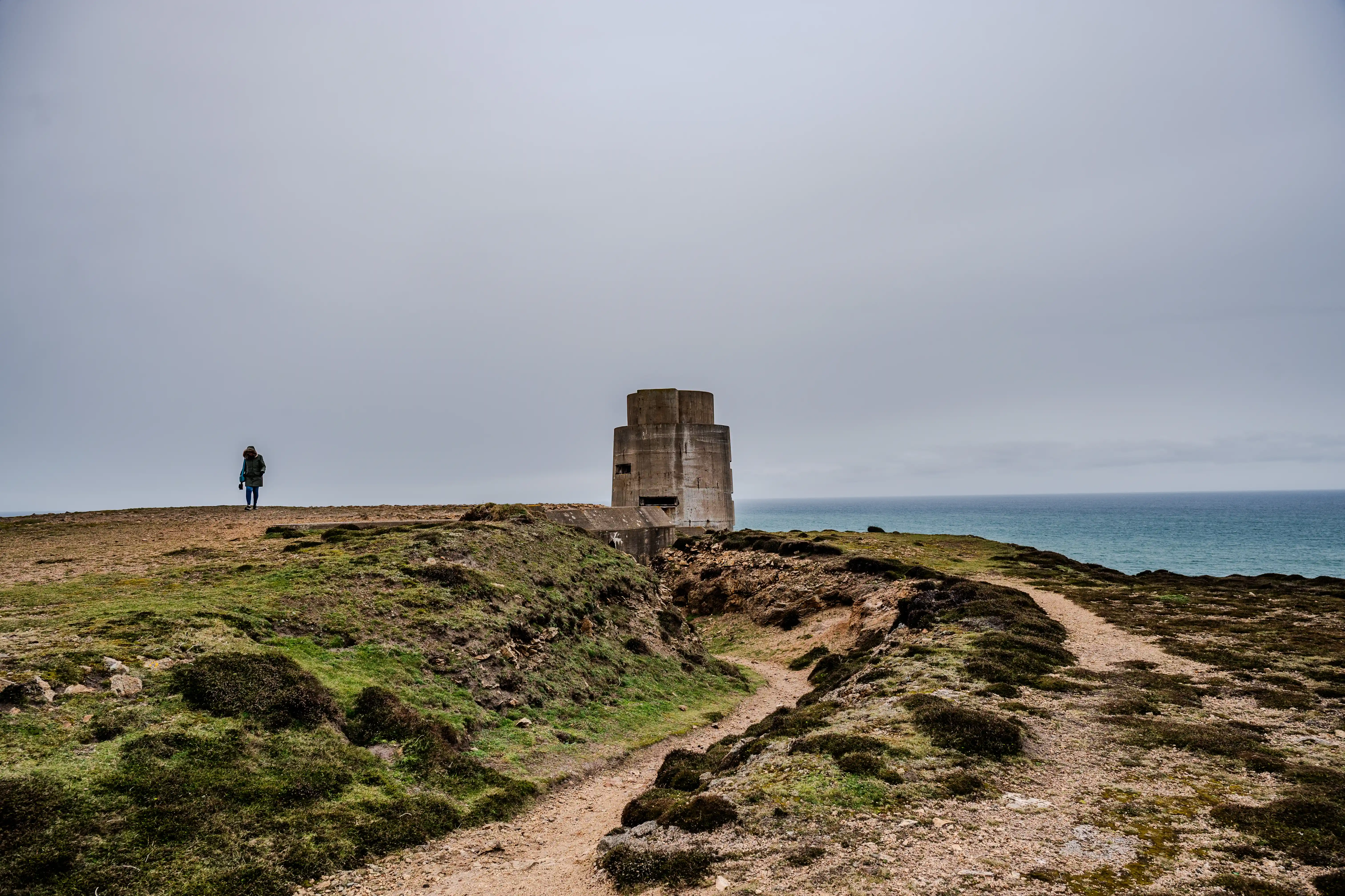  A person walks on a clifftop path towards a concrete bunker by the sea under a cloudy sky.