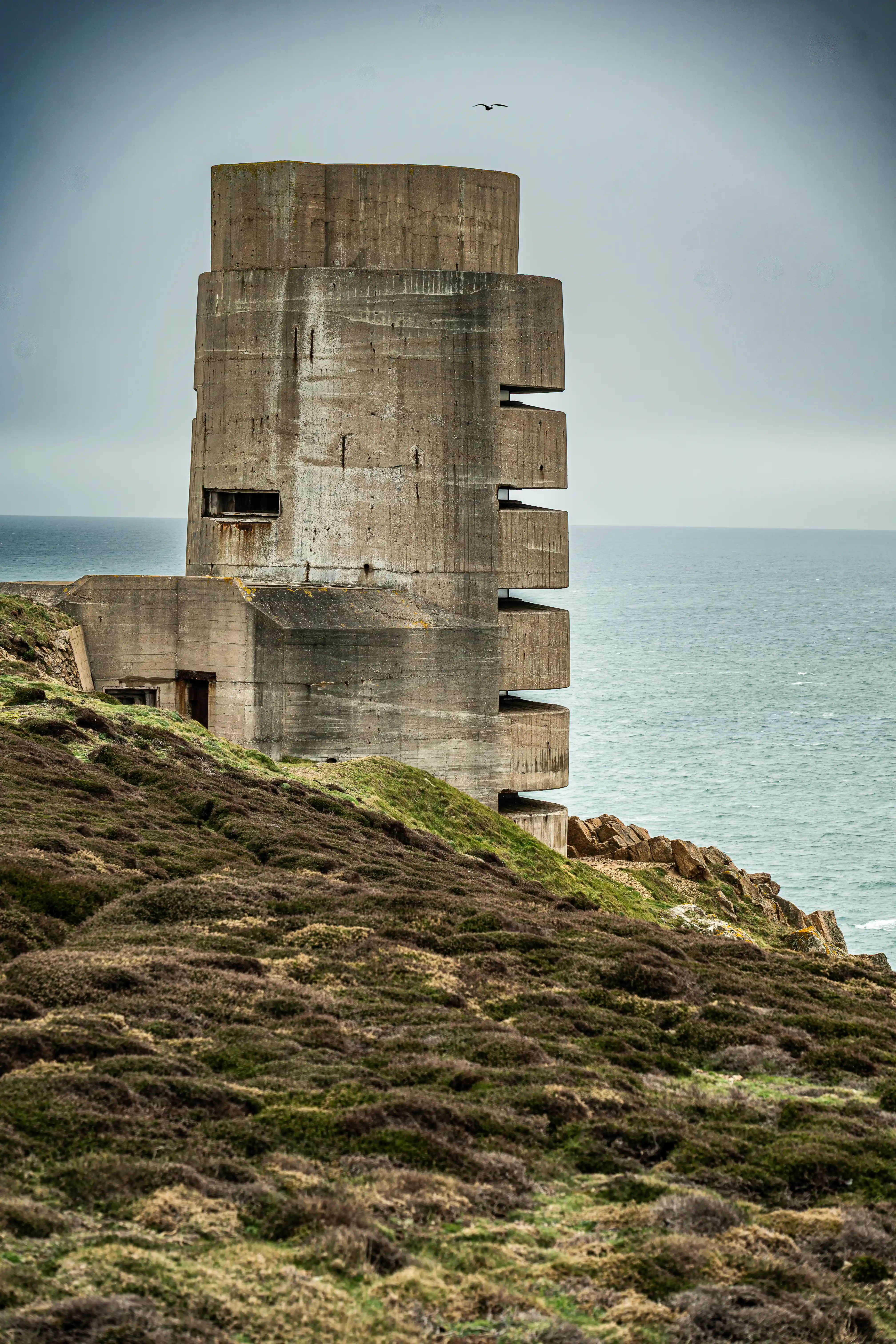A tall, multi-tiered concrete structure with narrow, horizontal openings stands on a grassy, heather-covered cliff edge. The sea is visible to the right under a cloudy sky, and a bird is in flight above.