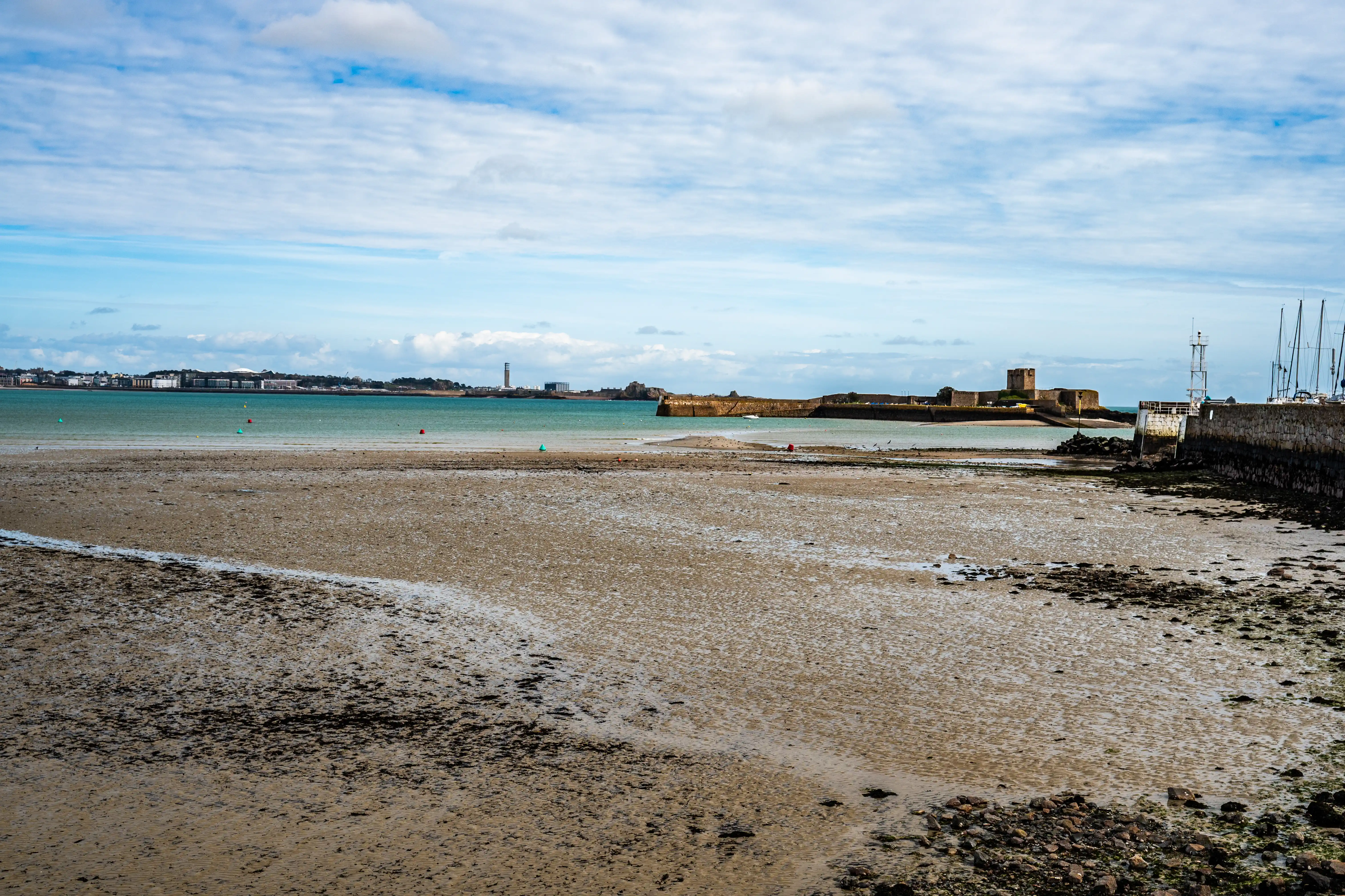 Wide shot of a beach at low tide with a stone castle in the distance and a harbour with yachts to the right.