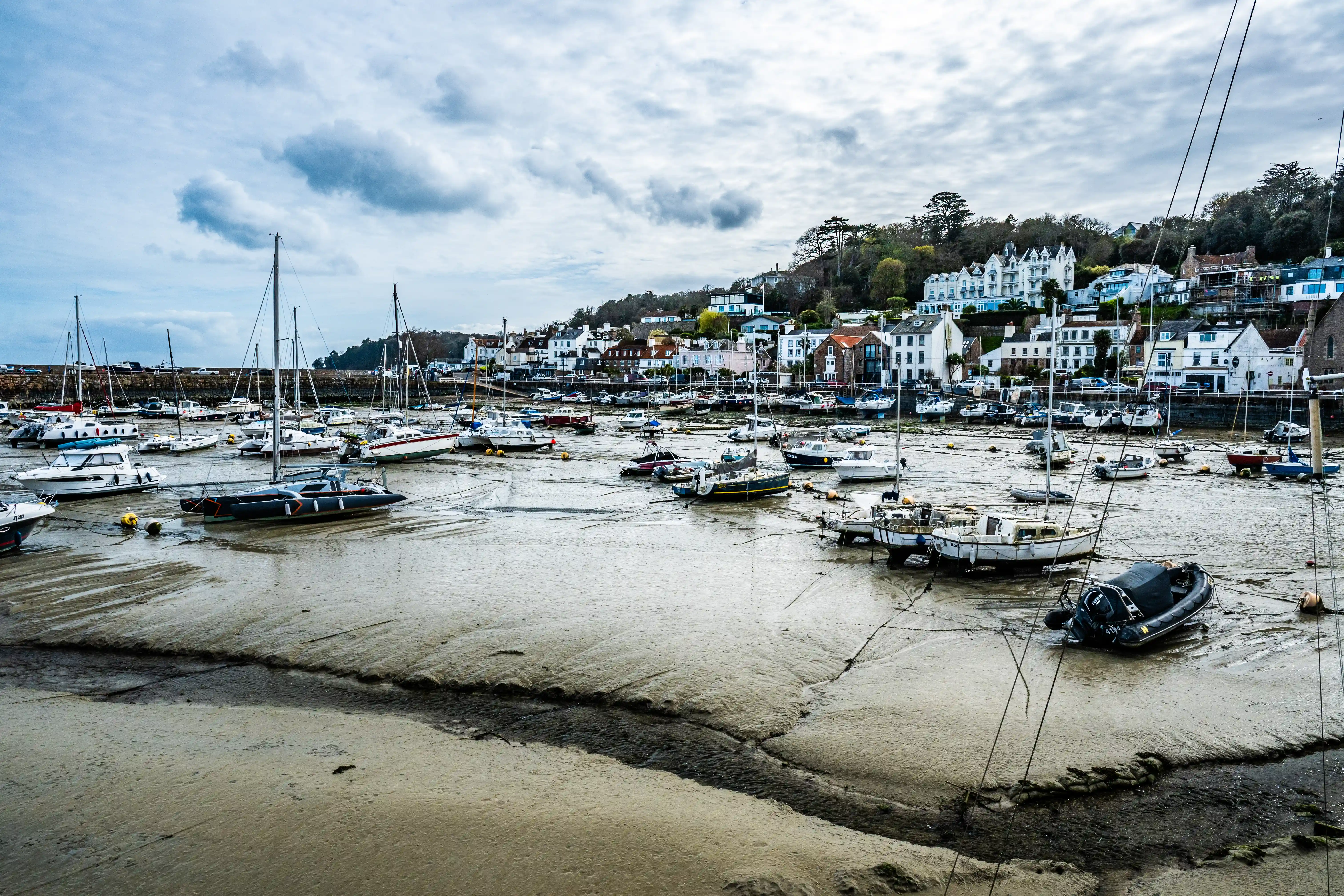 A harbour scene with many boats grounded on a muddy seabed due to low tide. Buildings are visible on a hill in the background under a dramatic sky.
