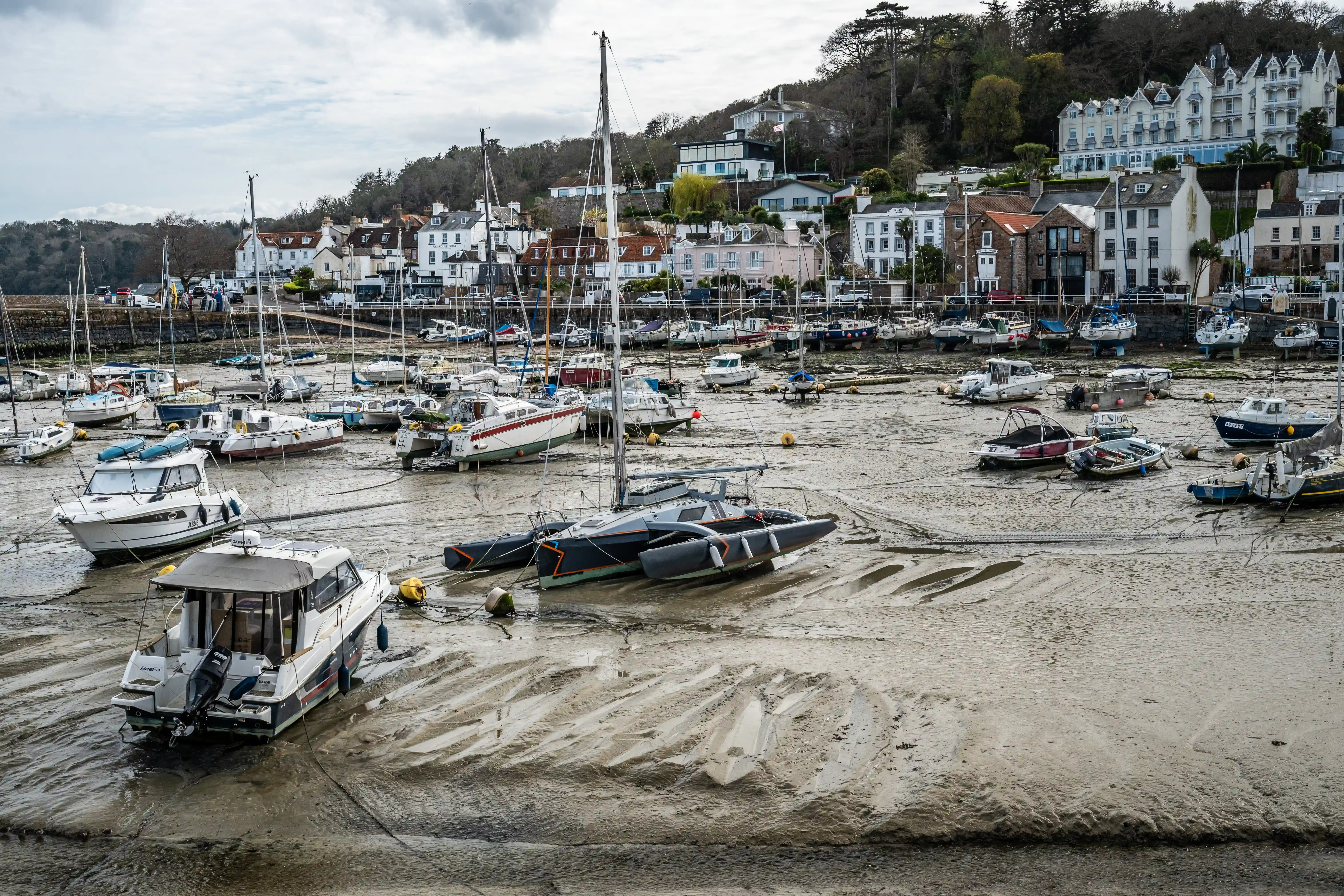 Several boats of different sizes, including yachts and motorboats, are resting on the wet, textured mud of a harbour floor at low tide. A coastal town with white and pastel-coloured buildings is visible on the hillside in the background, under an overcast sky.