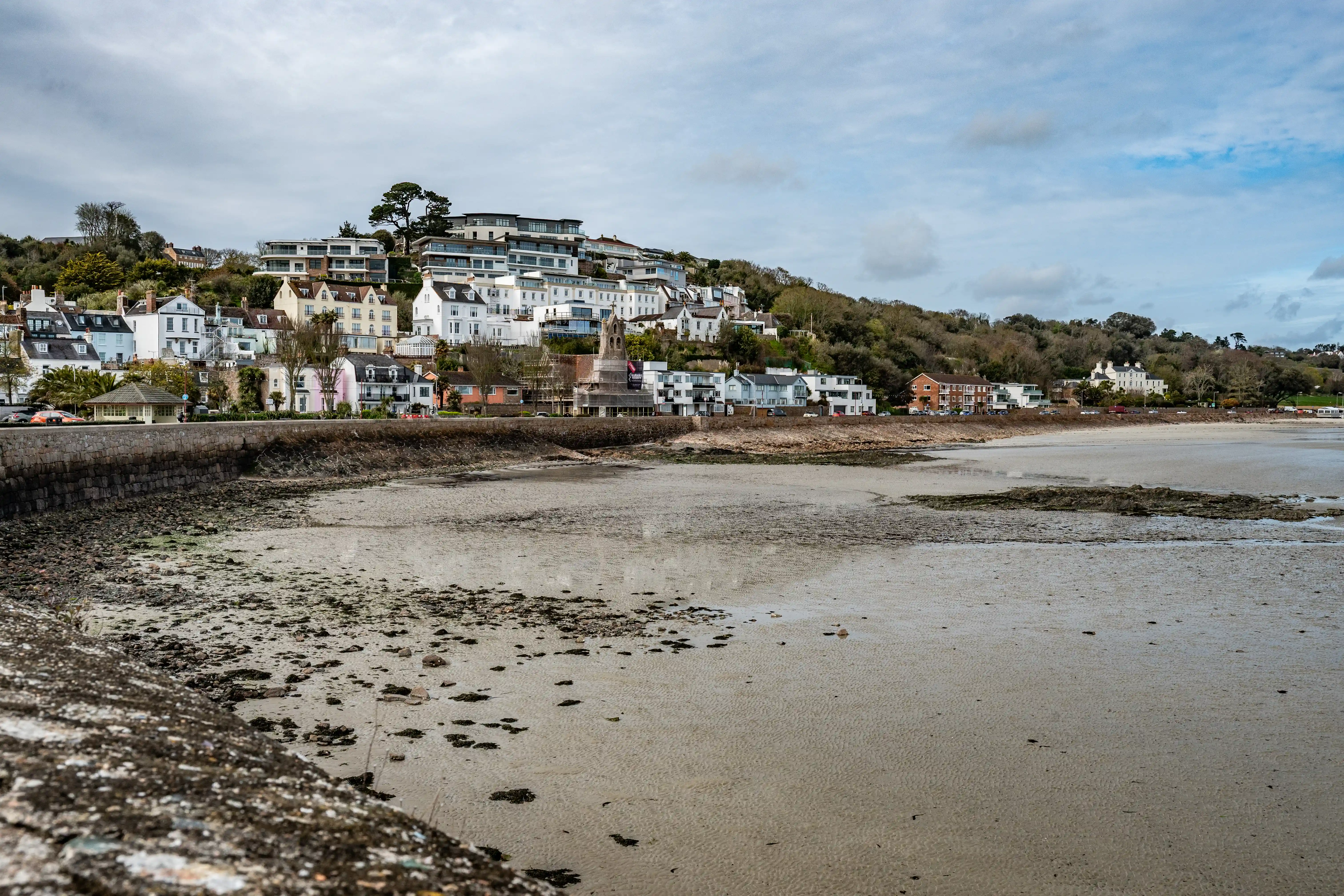 A seaside town with a mix of old and new buildings on a hill, with a beach exposed at low tide in the foreground under a cloudy sky.