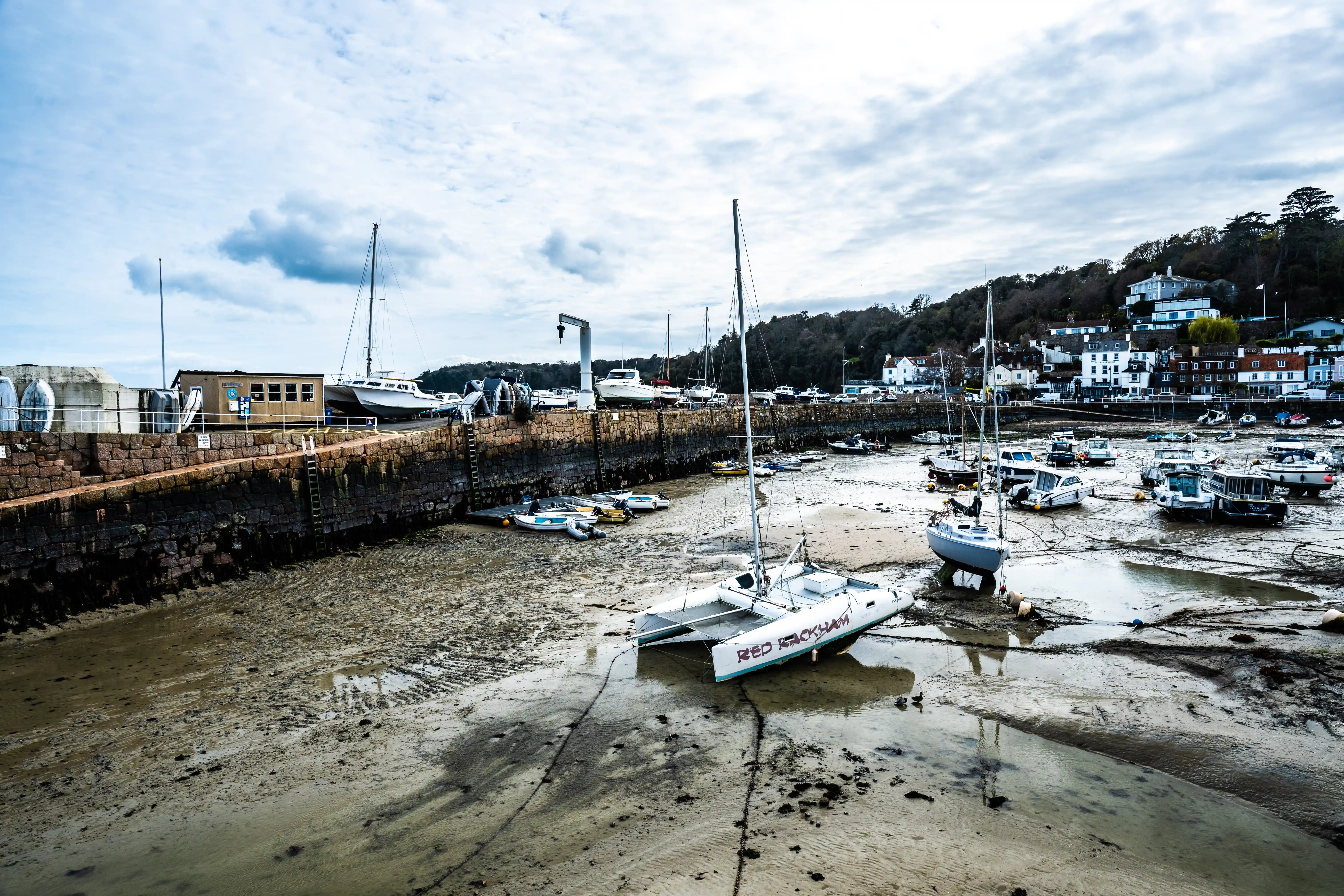Several boats, including a white catamaran, are beached on muddy ground during low tide in a harbour. A stone quay with buildings runs along the left, and a coastal town is visible on the hills in the background under a cloudy sky.