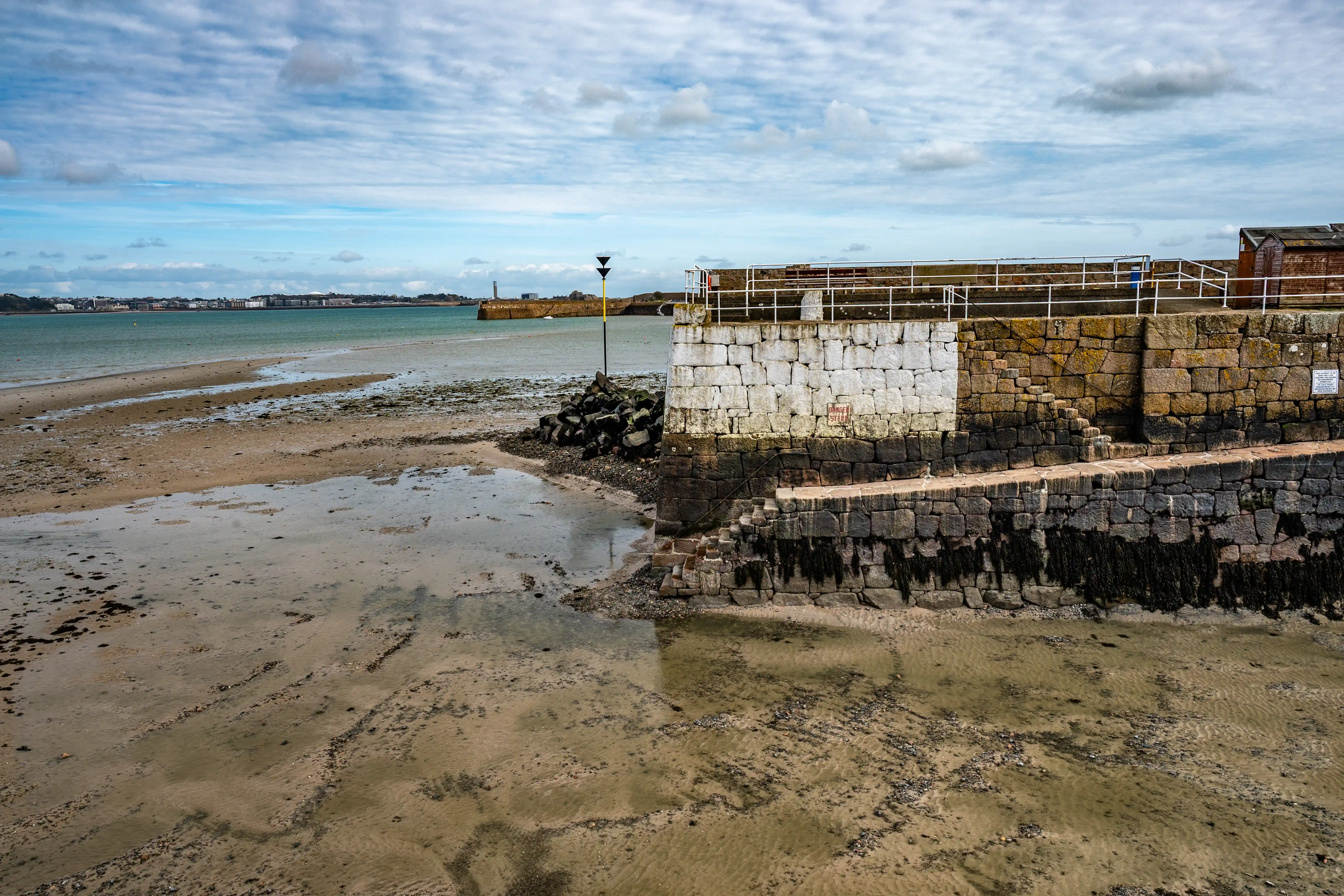 Wide shot of a seaside town's harbour at low tide, showing a sandy beach with shallow water and a large stone pier with white railings and steps, under a cloudy sky.