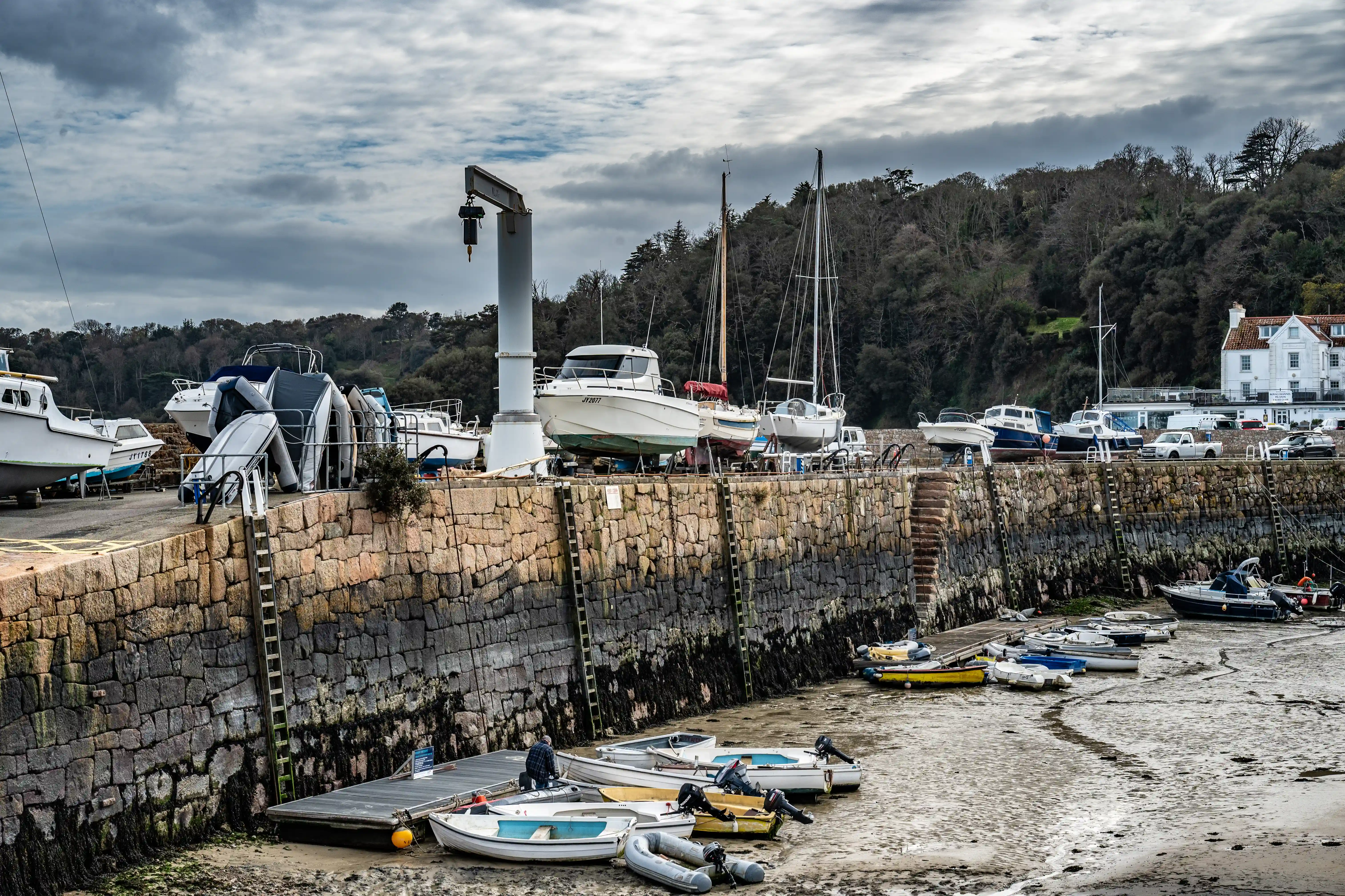 A harbour scene at low tide, with various boats on the muddy ground. A stone wall runs along the front, with a crane visible on the left and a white building on the hill in the background.
