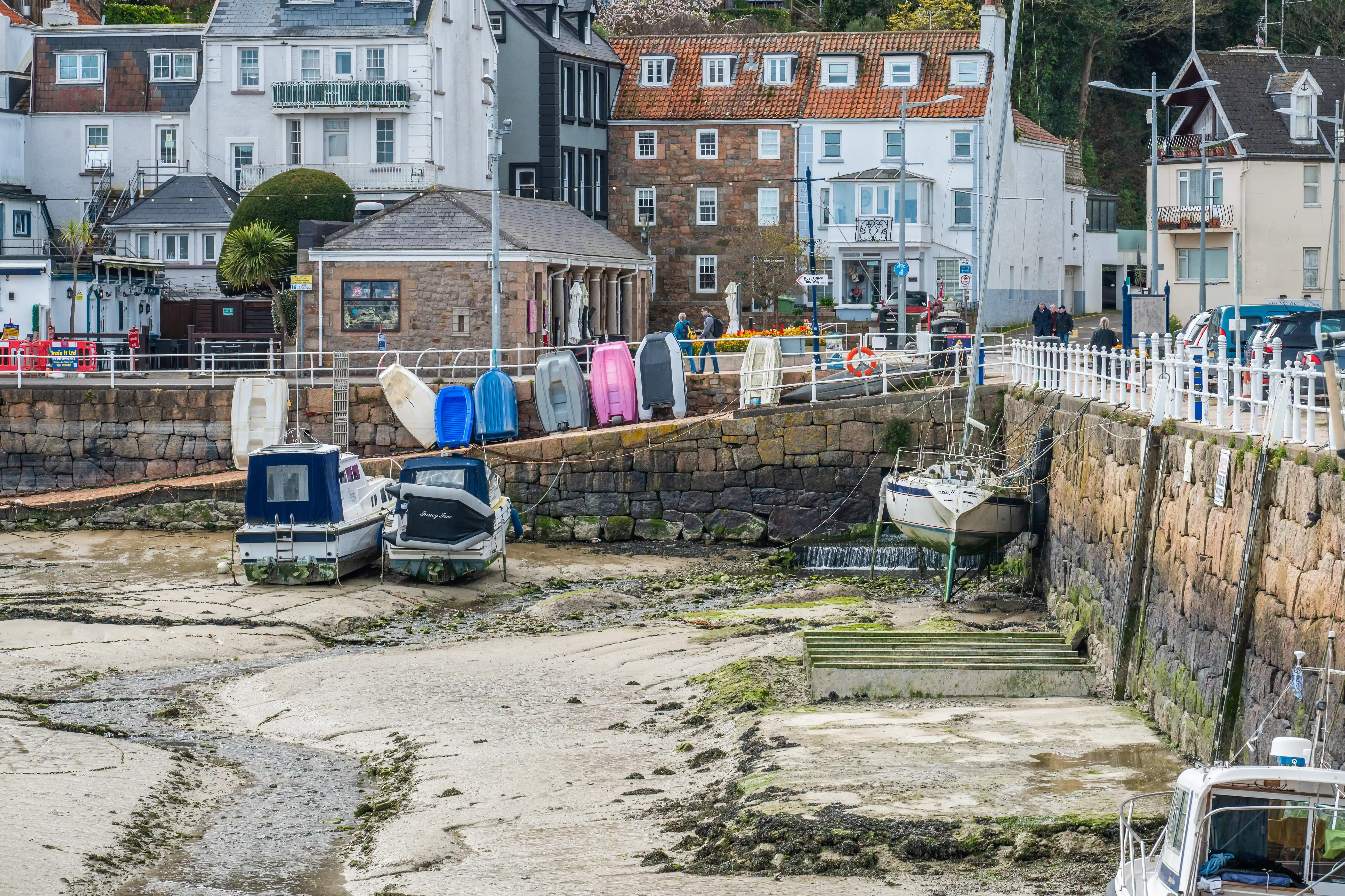 Several boats are moored along the stone walls of the harbor, which is mostly dry due to the low tide. Colorful kayaks or small boats are stacked on the harbor wall. Behind the harbor, there are various buildings, including houses and possibly shops or cafes, with a mix of architectural styles.