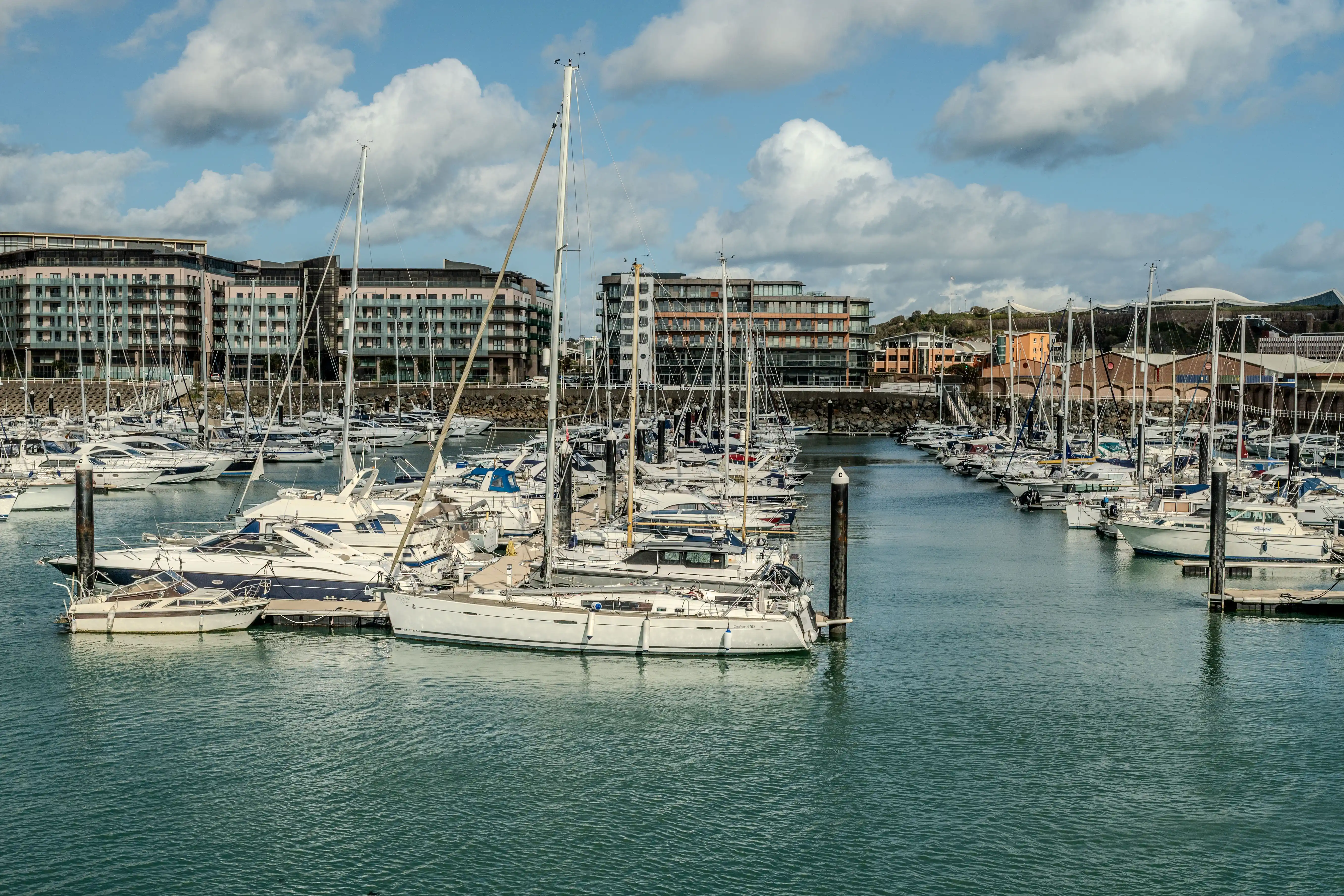 A wide shot of a marina filled with numerous sailboats and motor yachts, with modern apartment buildings and waterfront architecture in the background under a partly cloudy blue sky.