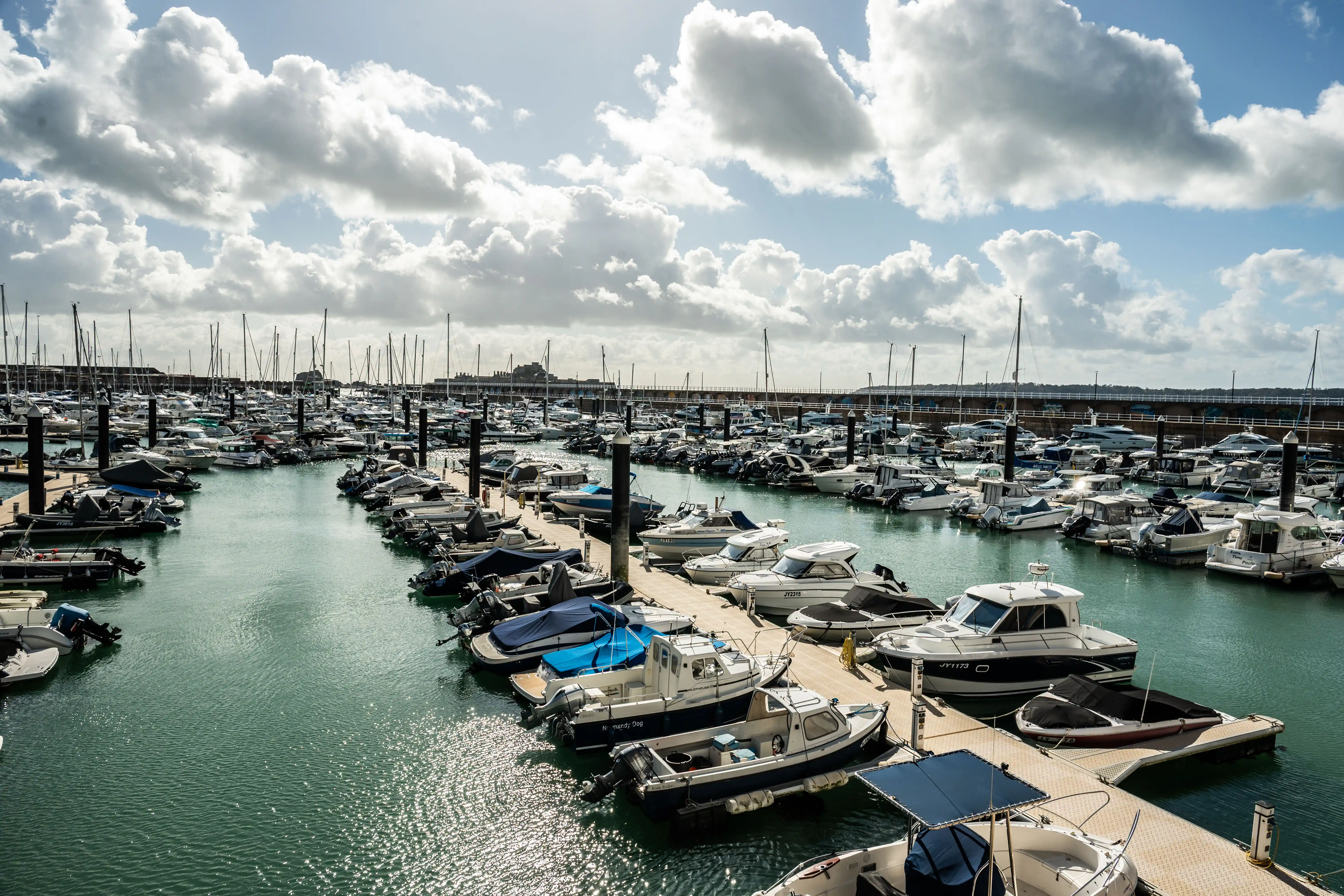 A busy marina filled with numerous boats moored along pontoons. The water is turquoise and reflects the sunlight. The sky is blue with dramatic white clouds, and a distant castle is visible on a hill.